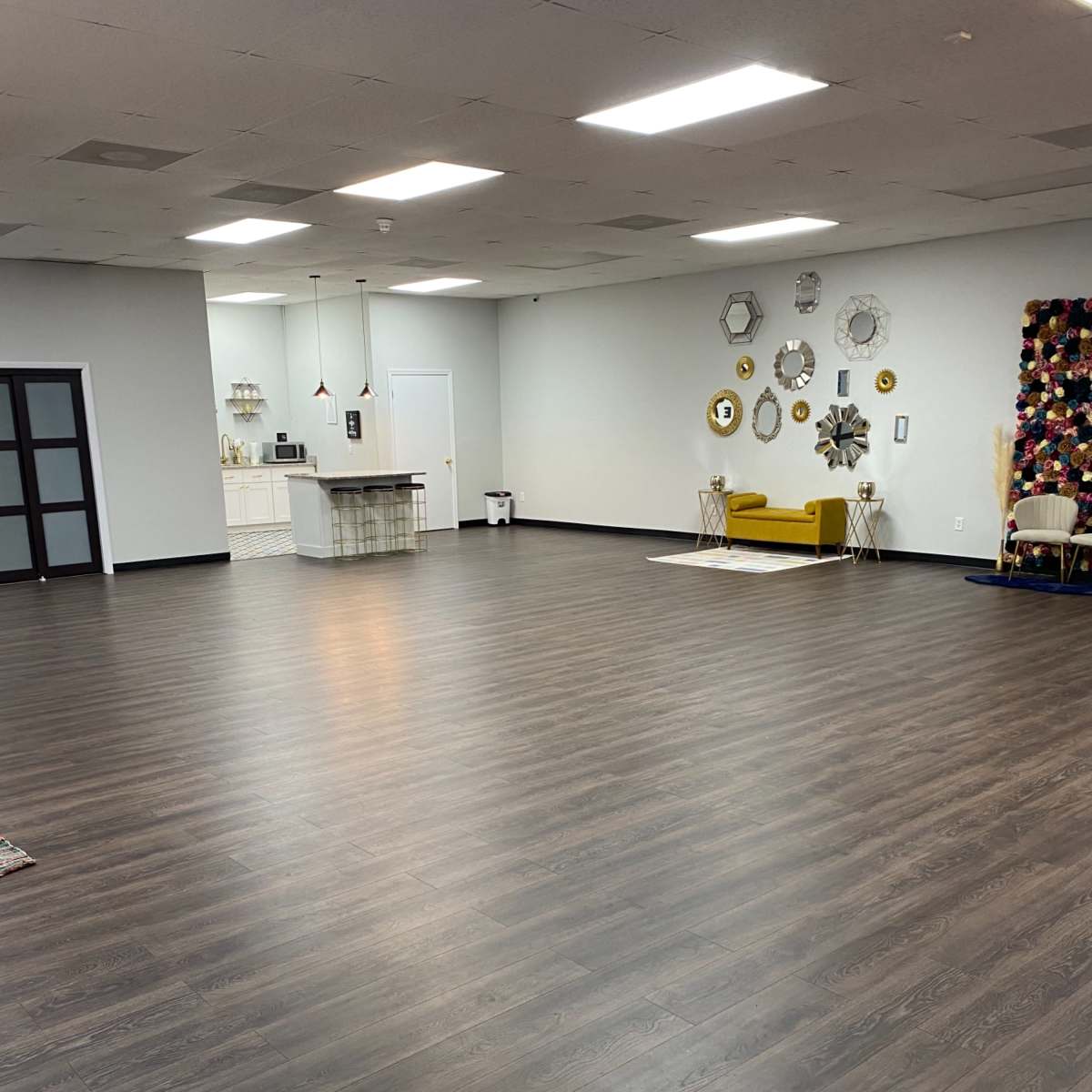 A spacious, empty room with dark wooden flooring, a kitchenette in the background, and decorative wall hangings on one side.