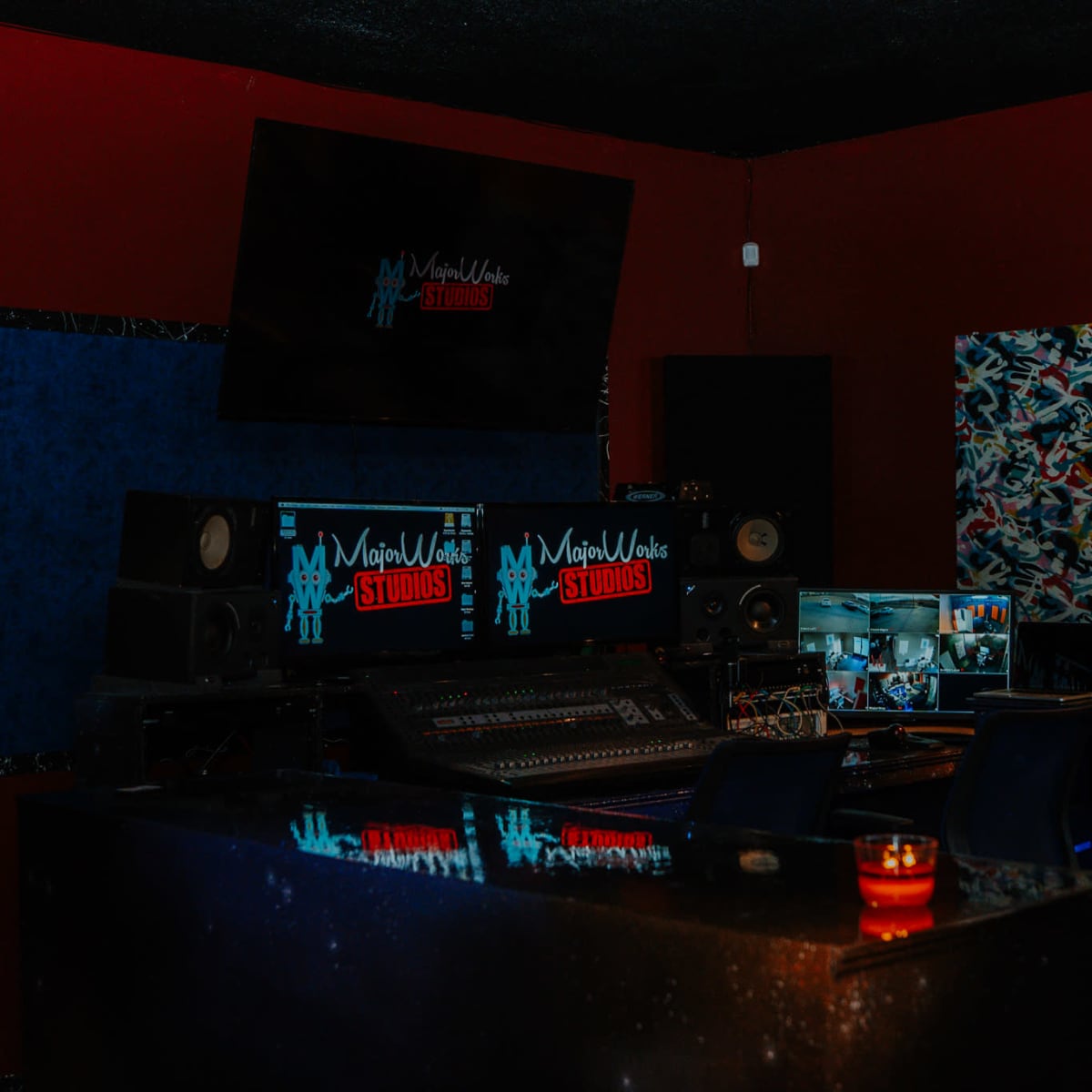 A dimly lit music studio with a control desk, multiple screens, and colorful wall art.