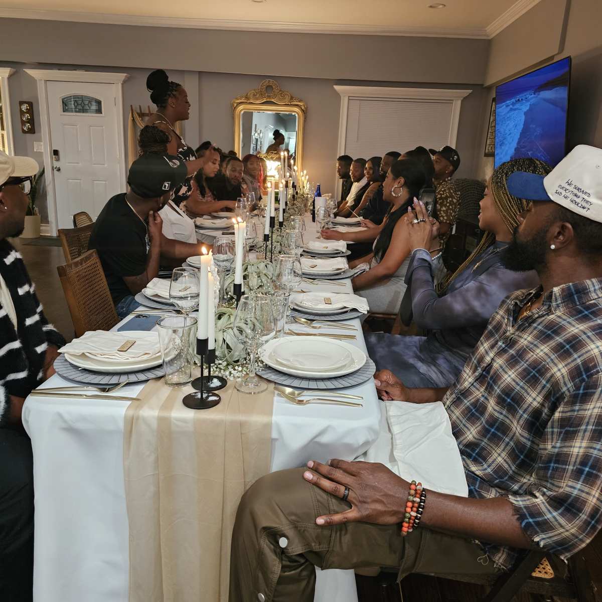 A long dining table is set with plates and candles, populated by guests engaged in conversation during a gathering in a well-decorated room.