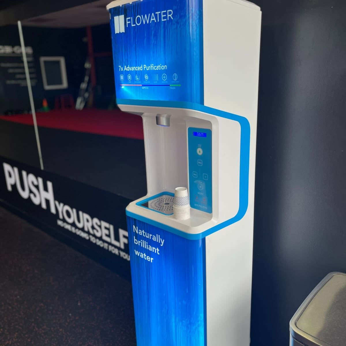 A water purification station with a sleek design, labeled "FLOWATER," featuring a button panel and a compartment for cups.