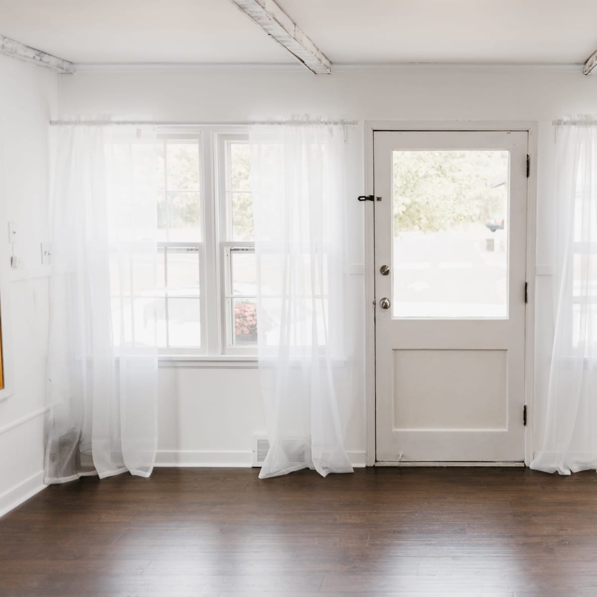 A bright, minimalist room features two windows with sheer white curtains and a white door leading outside.