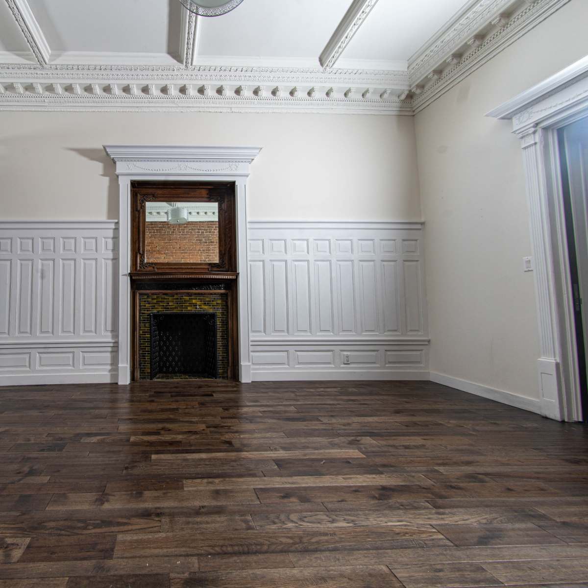A spacious interior room with wooden flooring, wainscoted walls, and a fireplace with a mirror above it, all under a decorative ceiling.