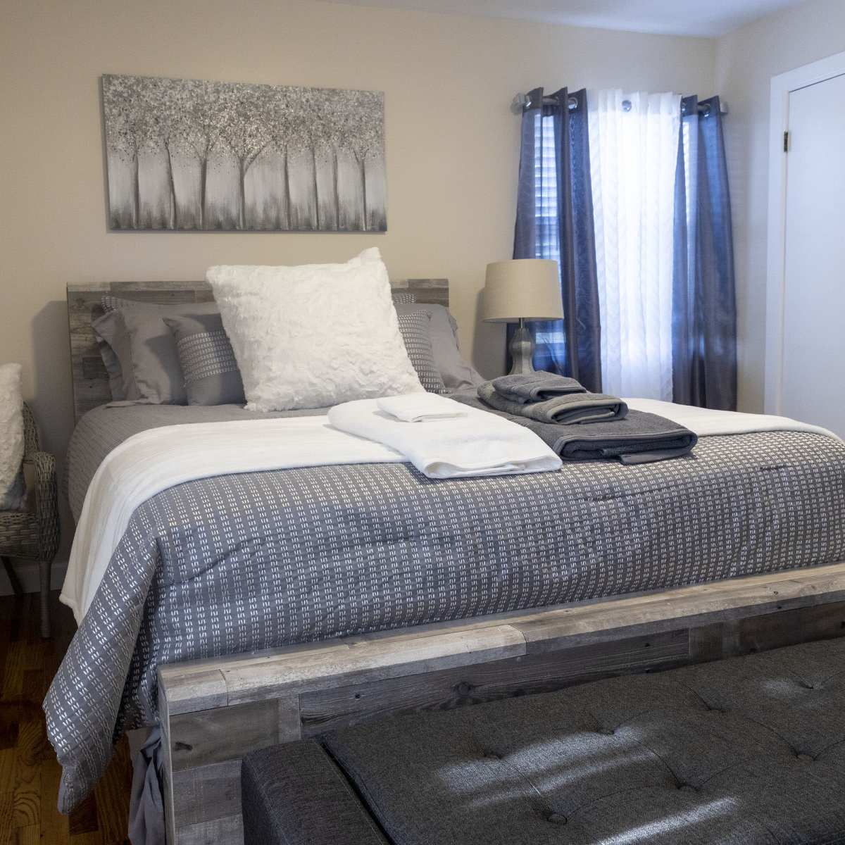 A neatly arranged bedroom with a wooden bed, gray bedding, a decorative wall art of trees, and a chair in the corner.