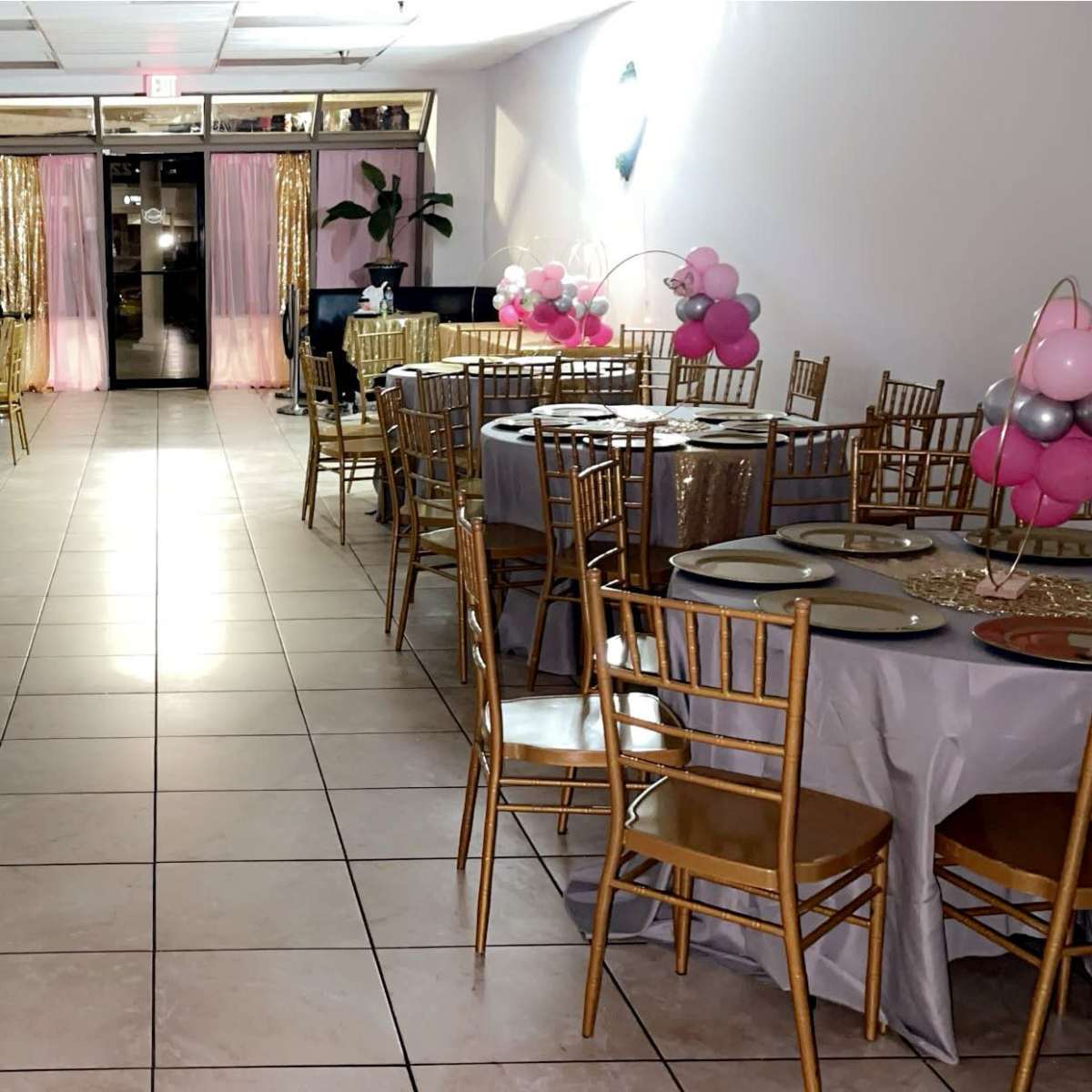 A banquet hall set up for an event, featuring round tables with gray tablecloths and gold chairs arranged in a line, adorned with pink and silver balloon decorations.
