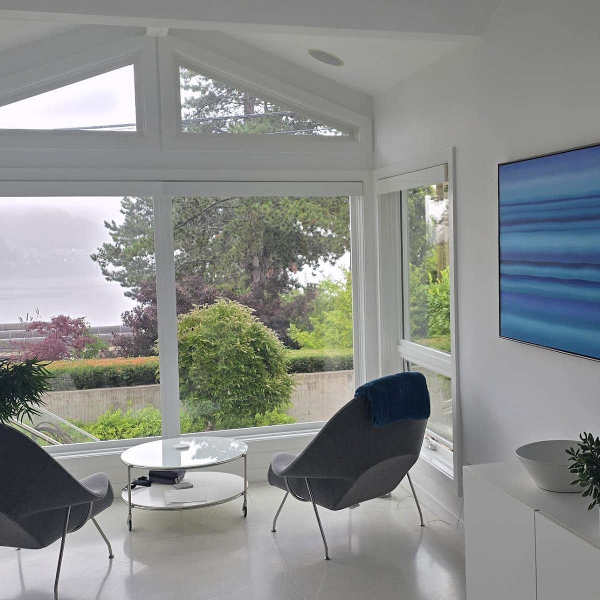 A brightly lit modern corner of a room features two gray chairs around a glass table, large windows offering a view of greenery, and a blue abstract painting on the wall.