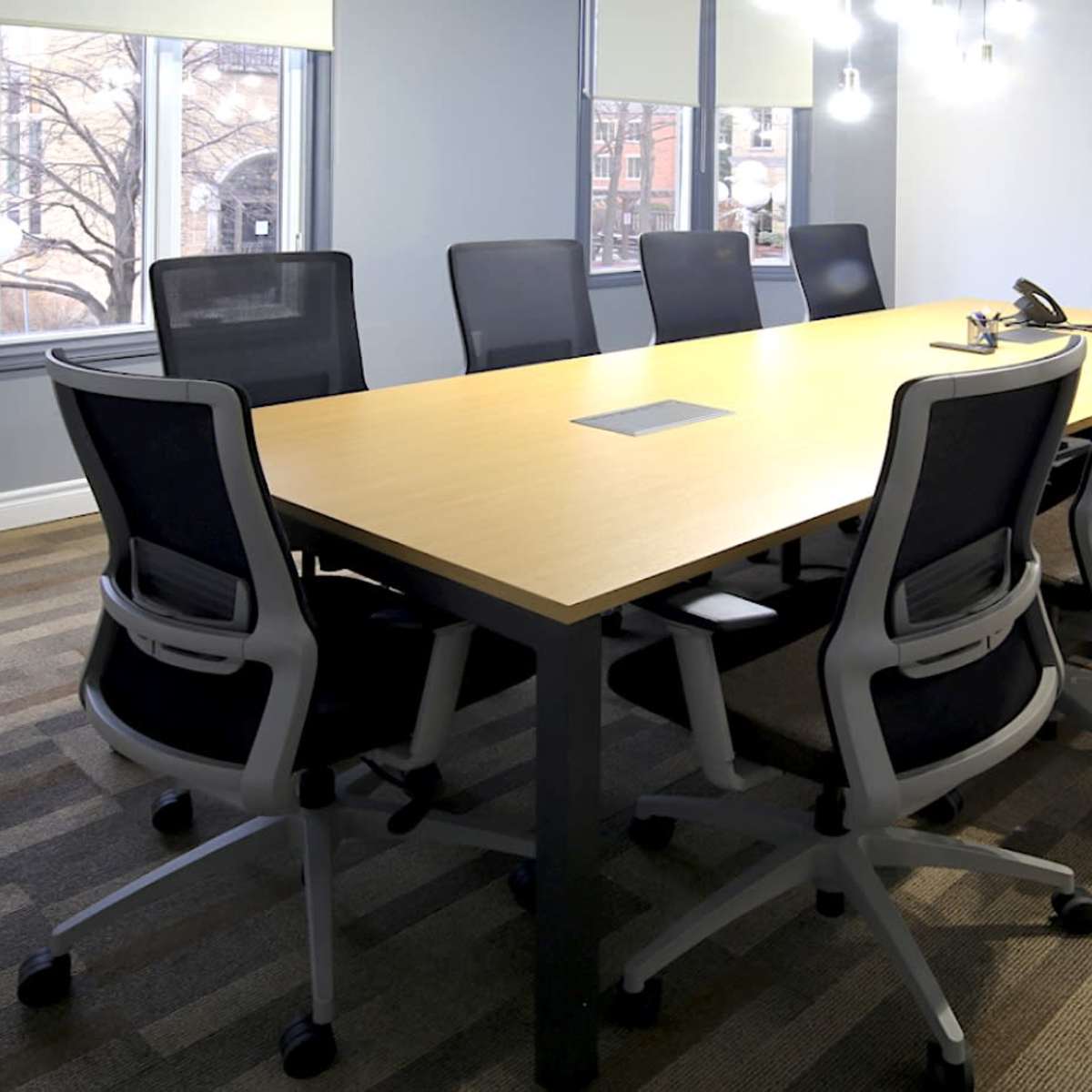 A modern conference room features a long wooden table surrounded by black ergonomic chairs, with large windows and a flat-screen television on the wall.