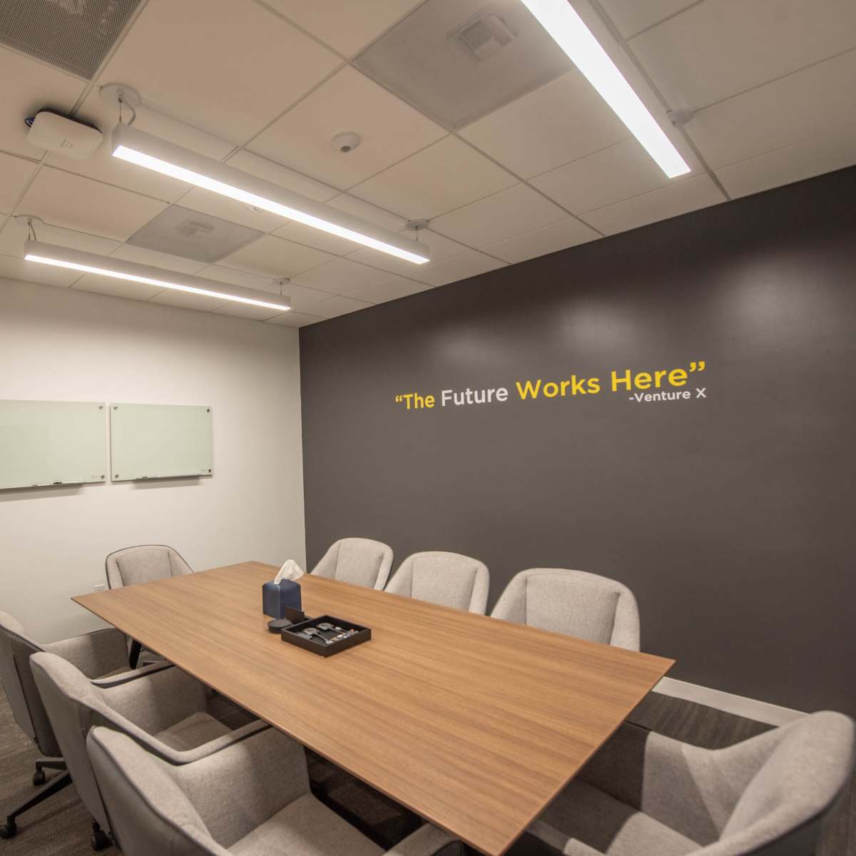 A modern conference room features a wooden table surrounded by six gray chairs and a wall with a motivational quote.
