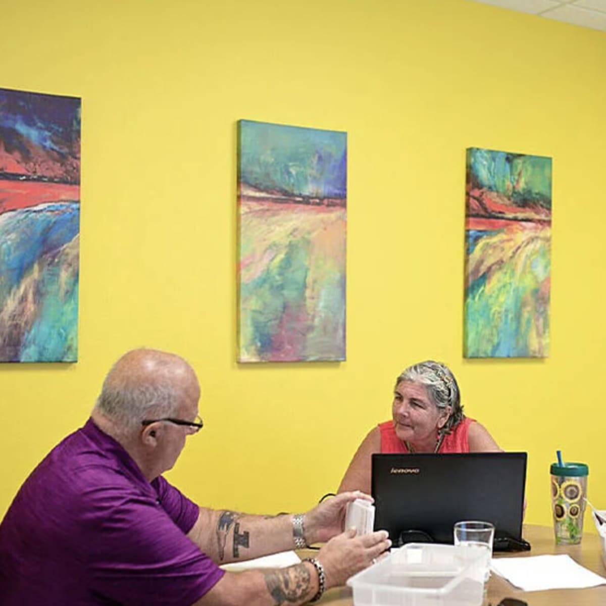 Two individuals are seated at a table in a brightly colored room, engaged in conversation, with artwork hanging on the wall and a laptop open in front of them.