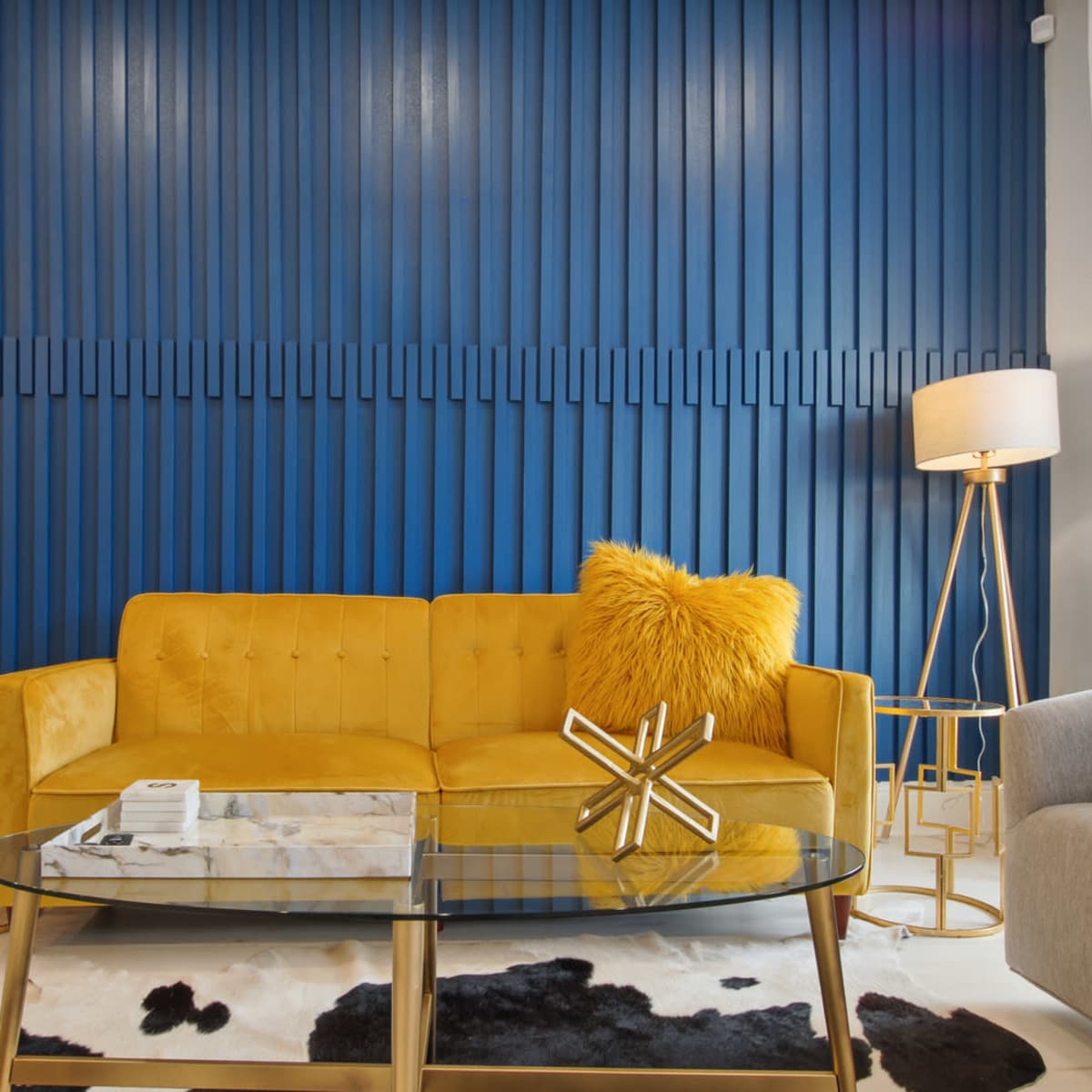 A yellow sofa with a fluffy pillow is positioned in front of a blue paneled wall, accompanied by a glass coffee table and a lamp, with a second lighter sofa nearby.