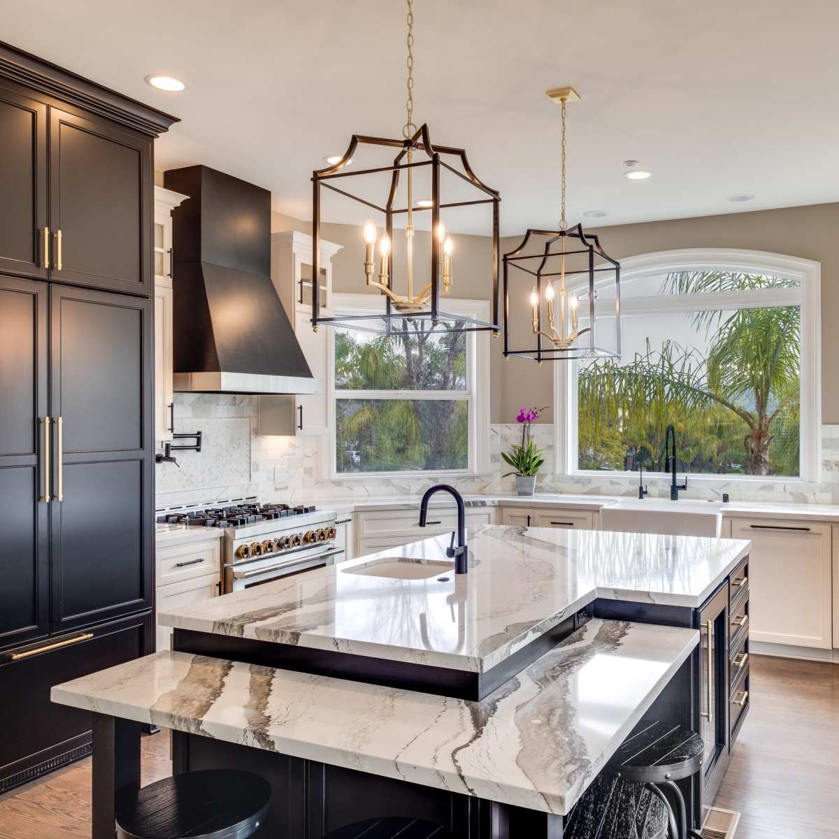 The kitchen features a large marble island, black cabinetry, and a window overlooking palm trees.