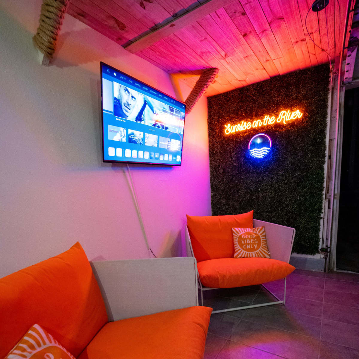 A cozy seating area with two orange chairs, a television mounted on the wall, and a glowing neon sign that reads "Sunrise on the River."