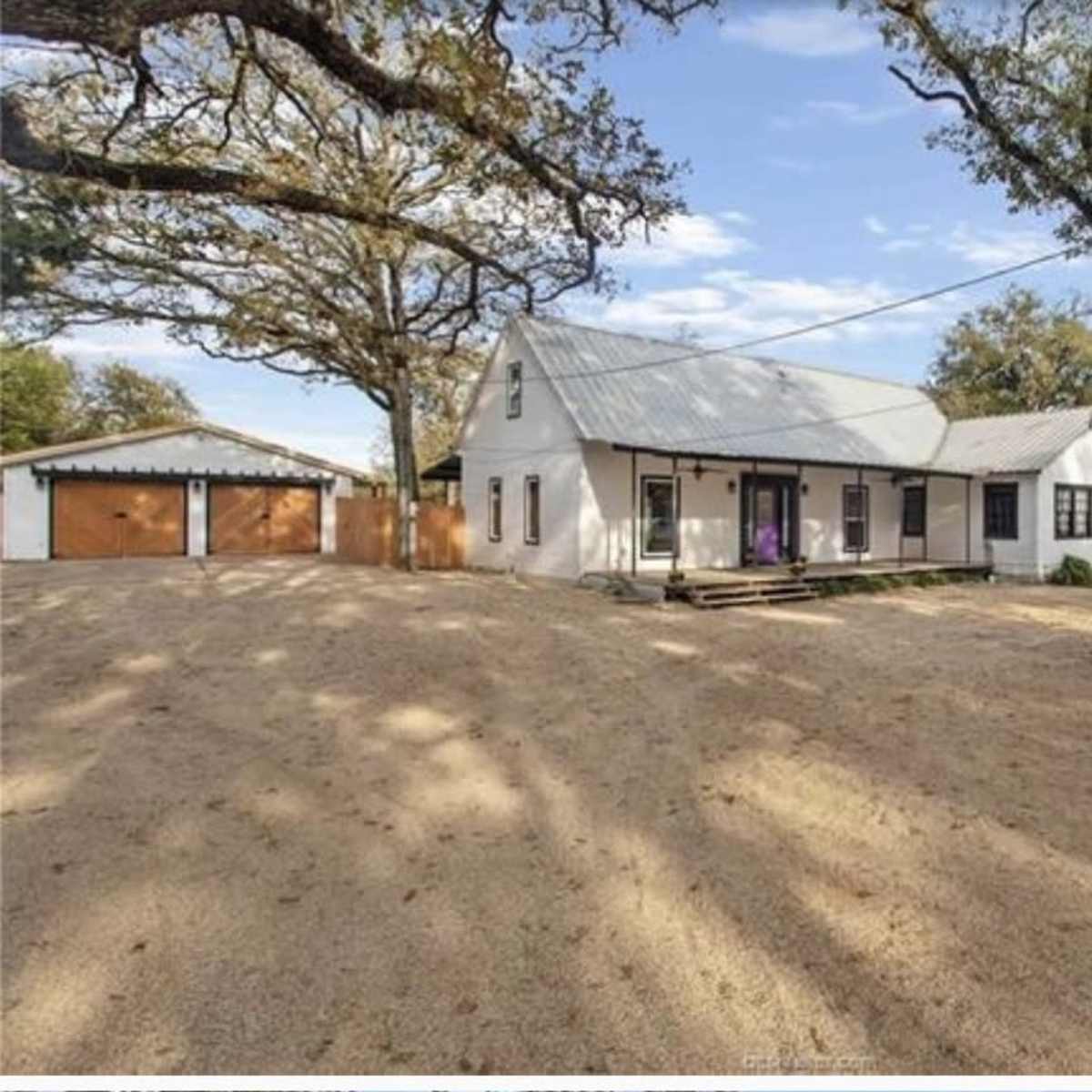 A white house with a metal roof, surrounded by gravel and trees, with a shed visible in the background.