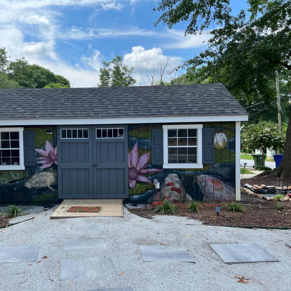 A garden shed with a mural of water lilies and koi fish is surrounded by a gravel pathway and landscaped greenery.