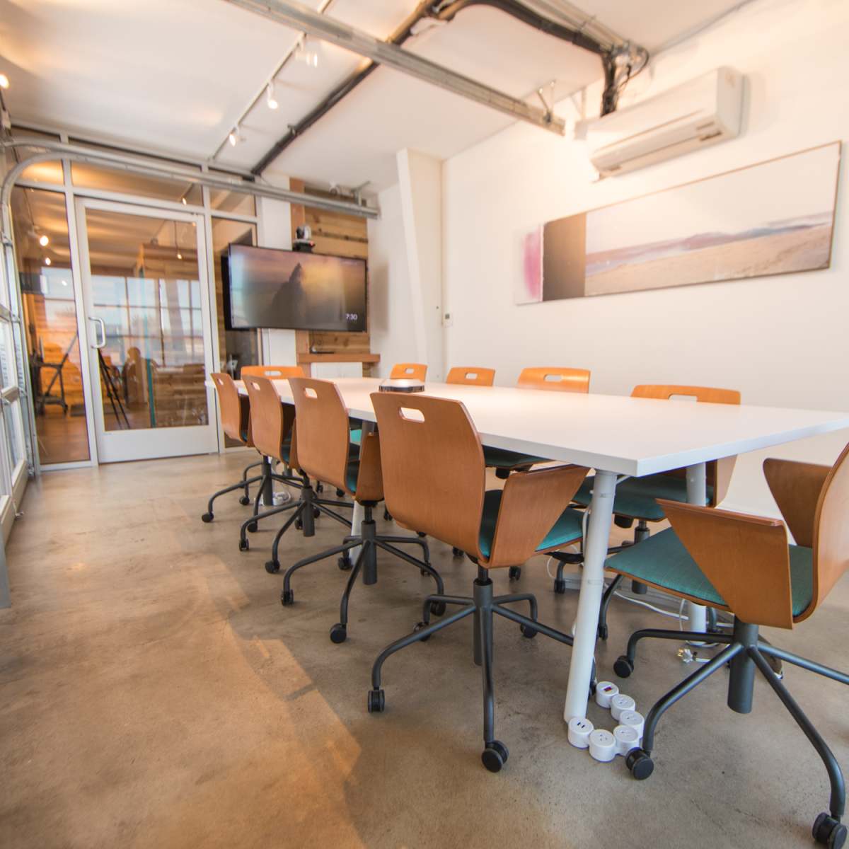 A modern conference room features a long white table surrounded by ergonomic wooden chairs and a large wall-mounted screen.