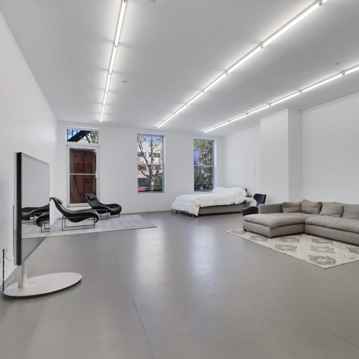 A spacious, modern room featuring a large sectional sofa, a bed, a TV on a stand, and two chairs, all against white walls and a concrete floor.