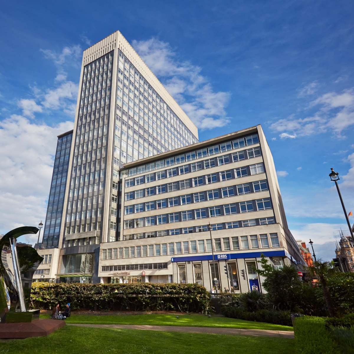 A modern high-rise building with a glass facade located next to a landscaped area with a large sculpture in the foreground.
