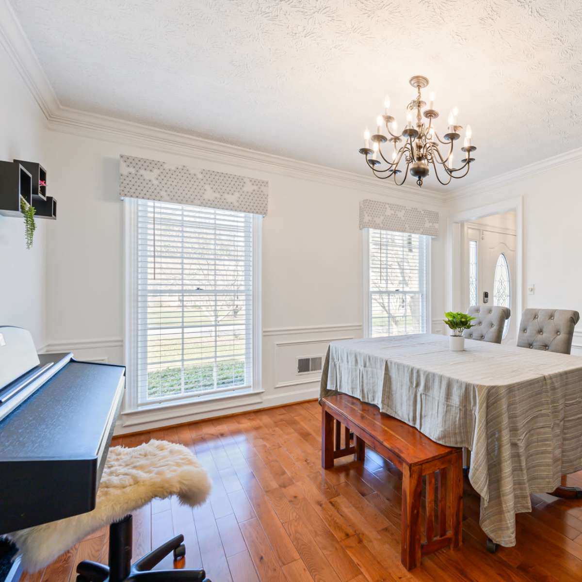 A well-lit dining room features a central table, two upholstered chairs, and large windows with white blinds.