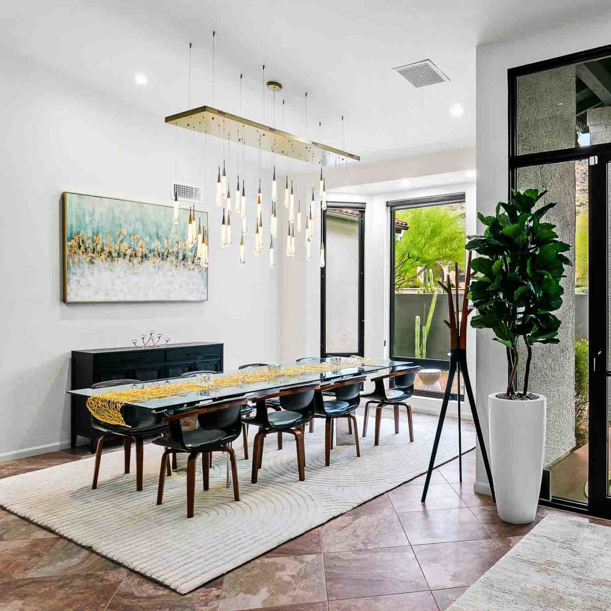A modern dining area features a long table surrounded by chairs, a decorative light fixture above, and large windows that open to an outdoor space.