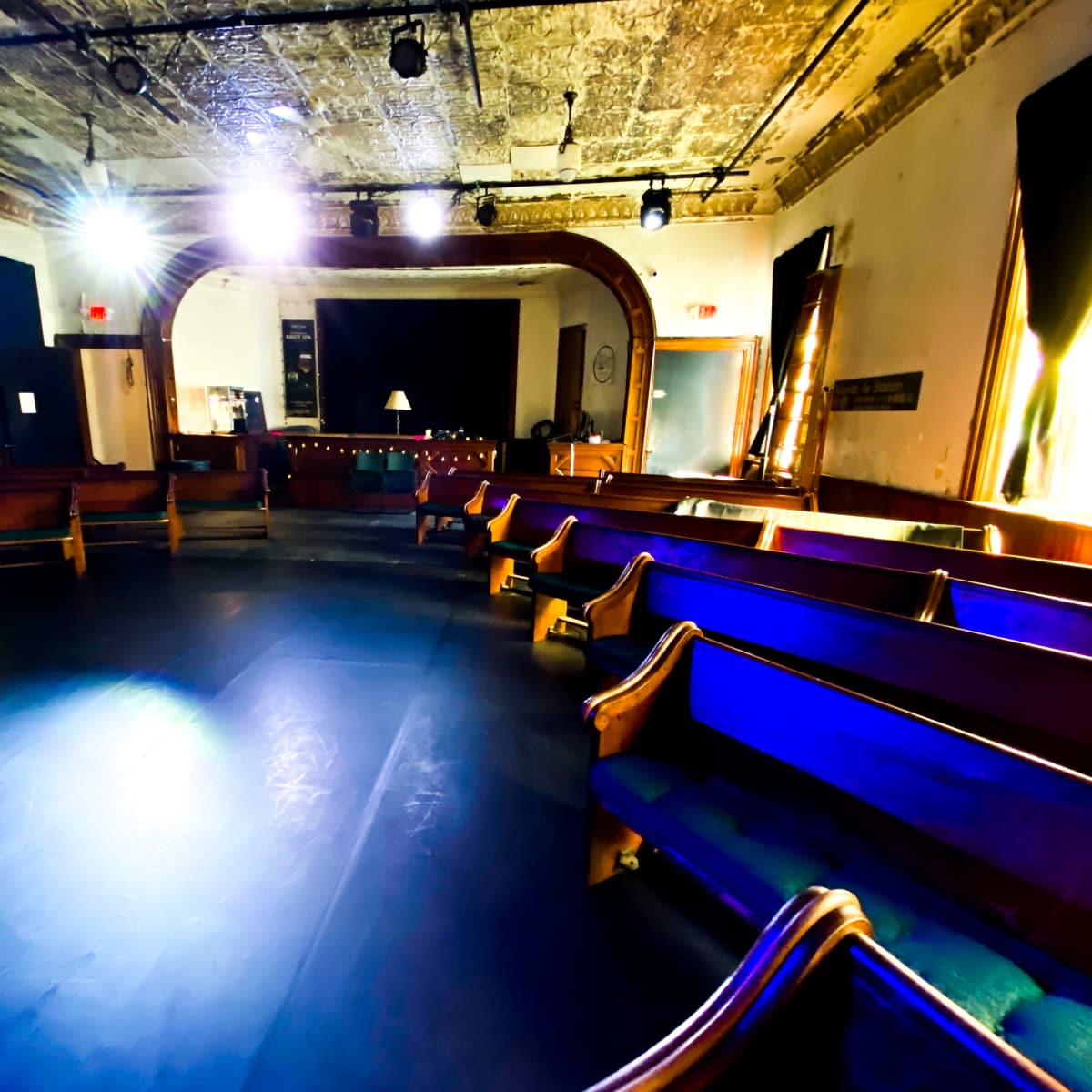 An empty, circular space with wooden benches arranged around a central area, illuminated by stage lights and featuring a backdrop with a sign reading "5TH STREET."