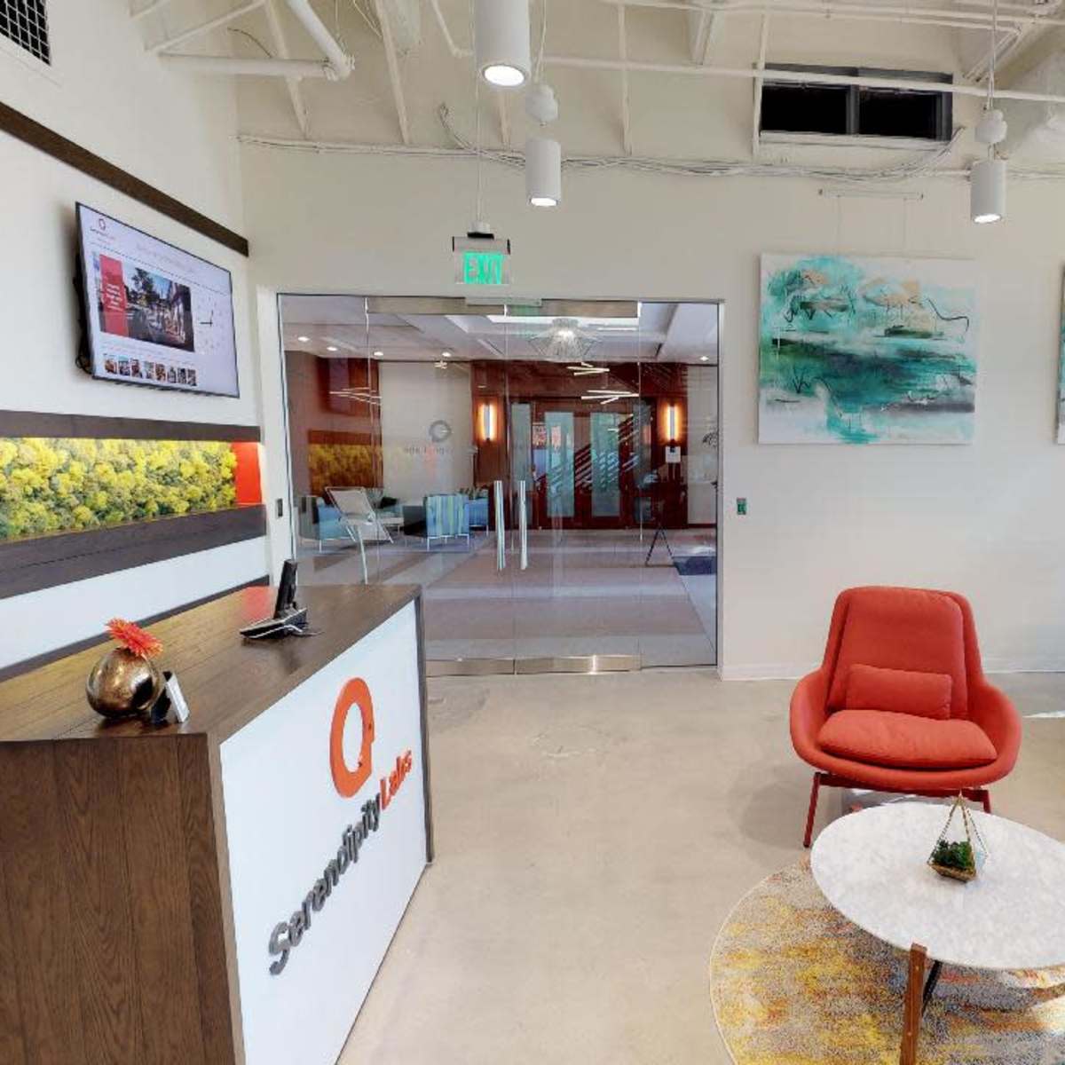 A modern office reception area featuring a sleek front desk, red chairs, a circular coffee table, and abstract artwork on the walls.
