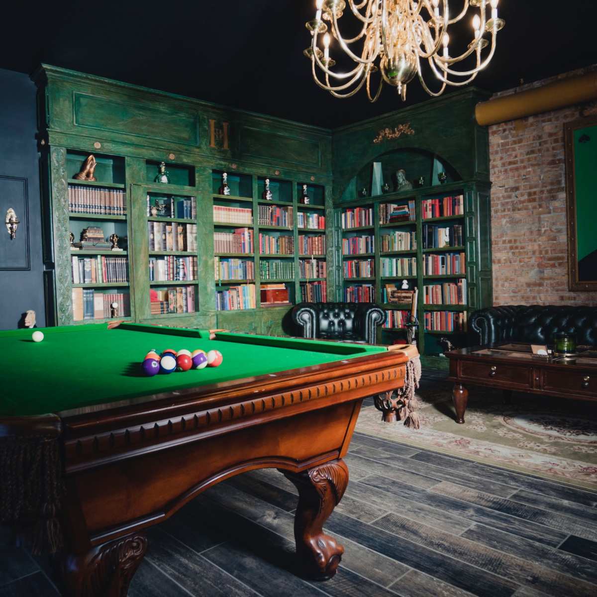 A room with a green pool table in the center, surrounded by bookshelves filled with books, a leather sofa, and a chandelier overhead.