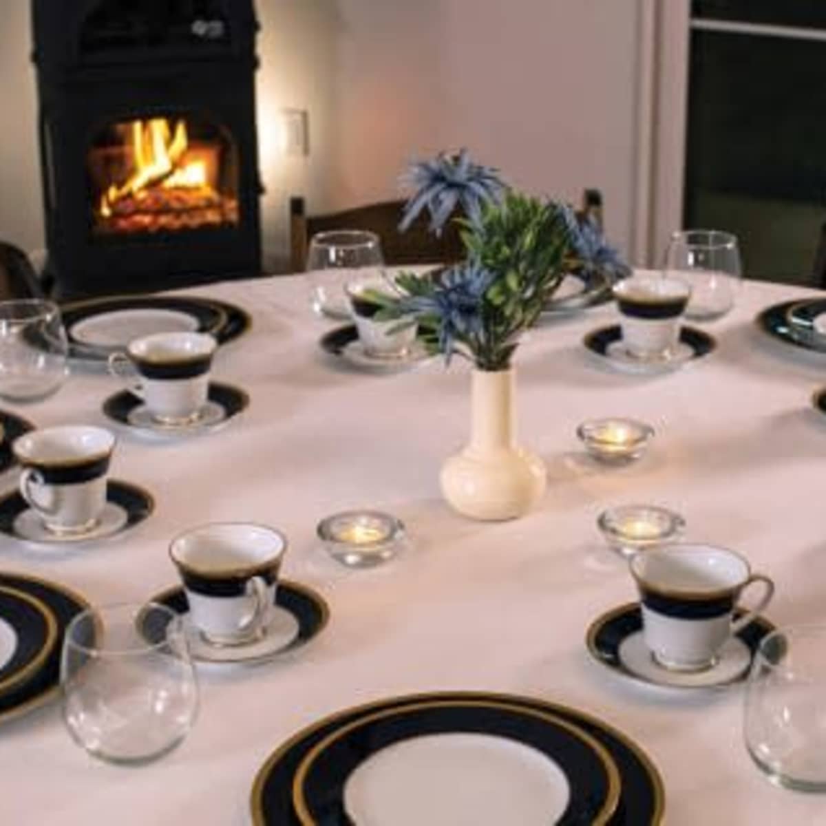 A round dining table is set with plates, cups, and glasses, centered by a vase of flowers and illuminated by candles, with a fireplace visible in the background.