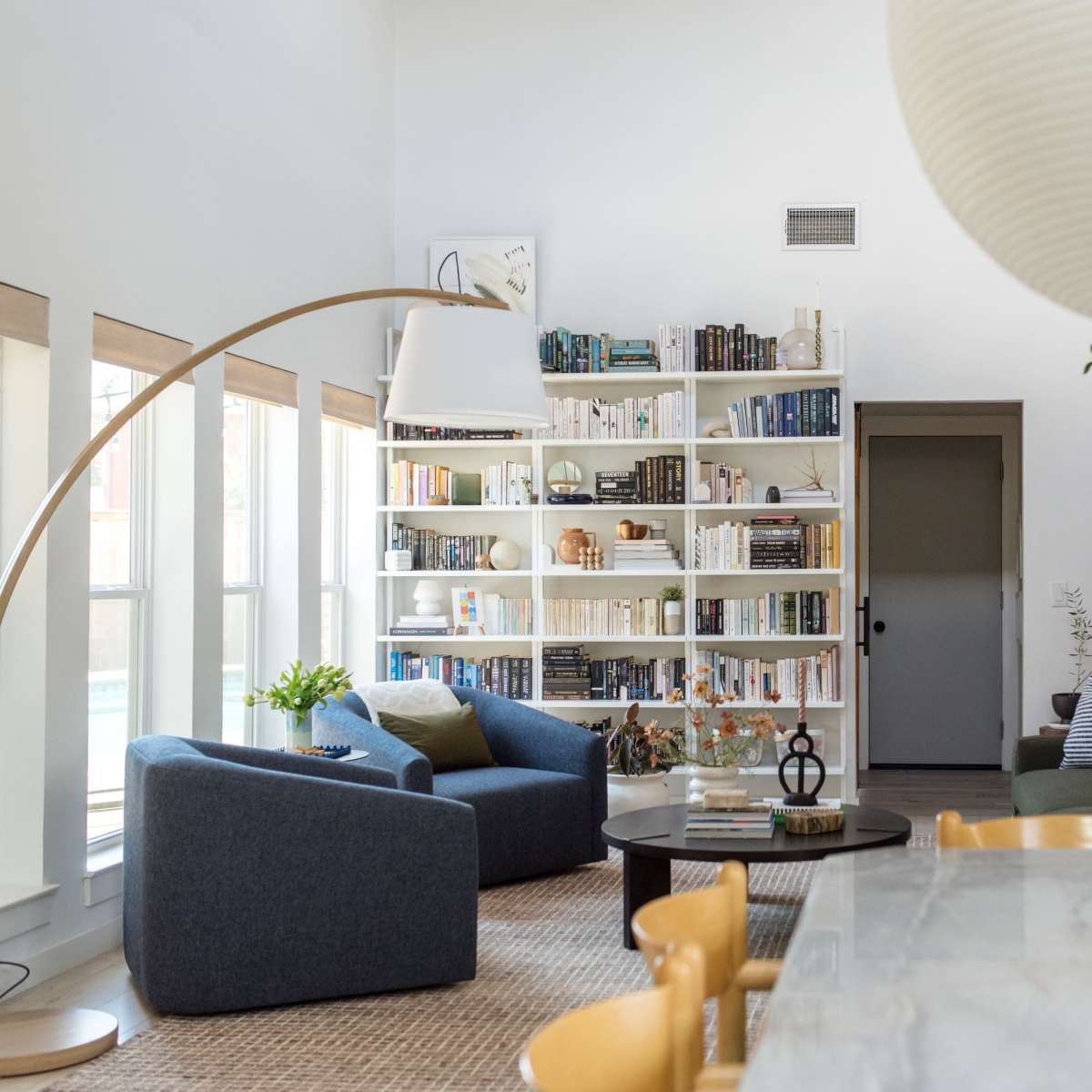 A modern living room with a large bookshelf, a marble dining table, two lounge chairs, and a staircase in a bright, airy space.