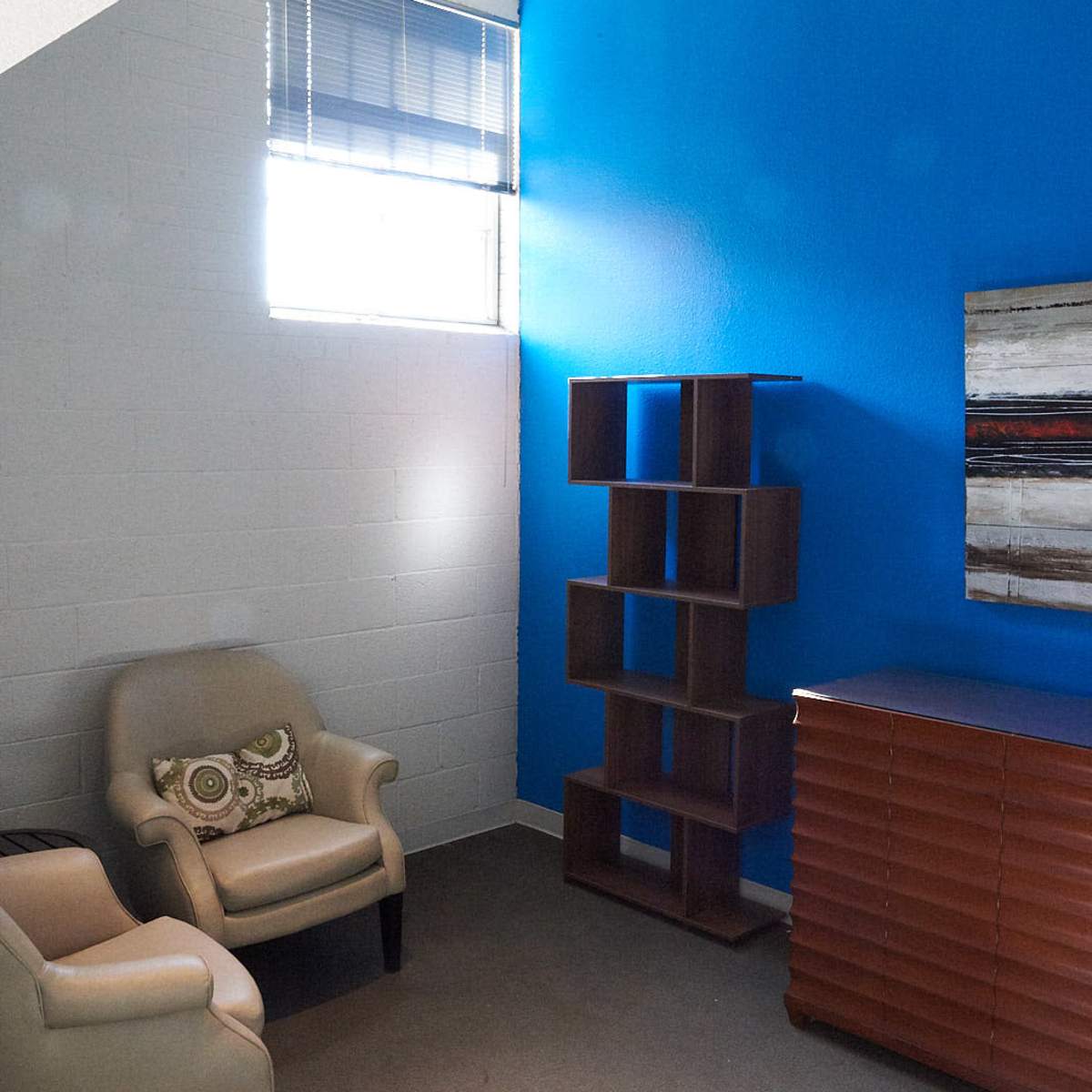 A small room with a blue accent wall, featuring a chair, a bookshelf, and a wooden dresser.