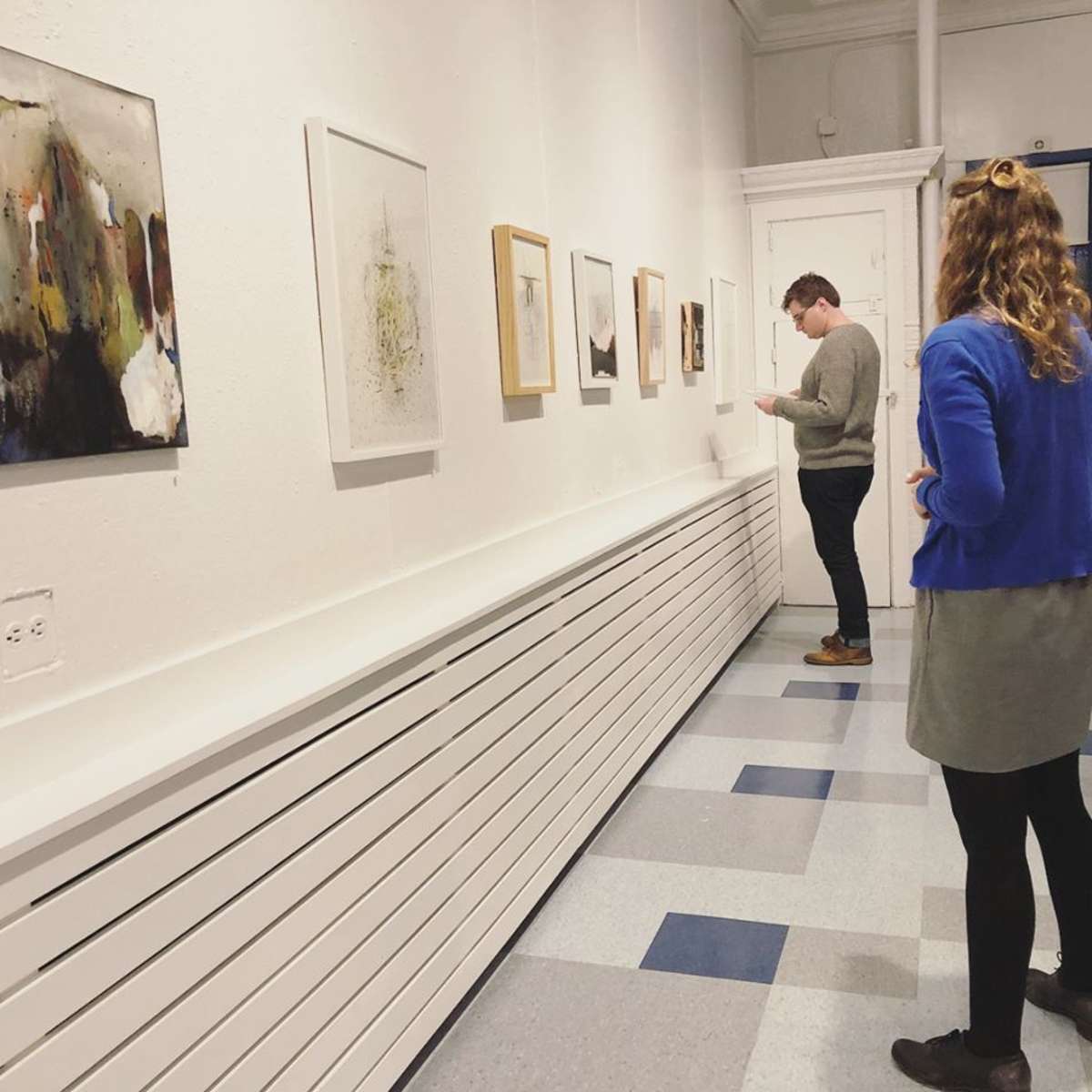 Two people are observing artworks displayed on a white wall in an art gallery.