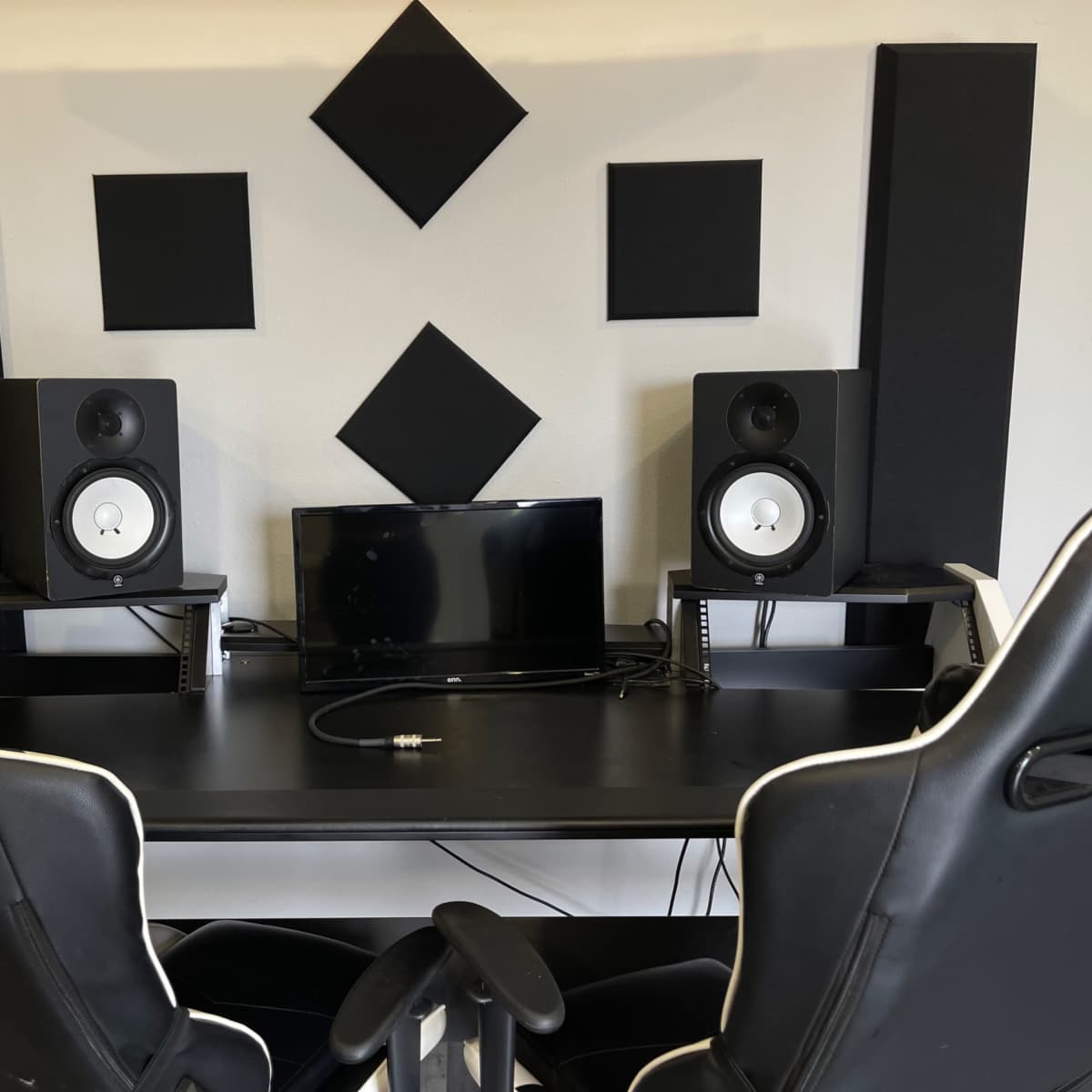 A modern workspace with dual black gaming chairs, a desk, two studio monitors, and a small television surrounded by soundproofing panels.