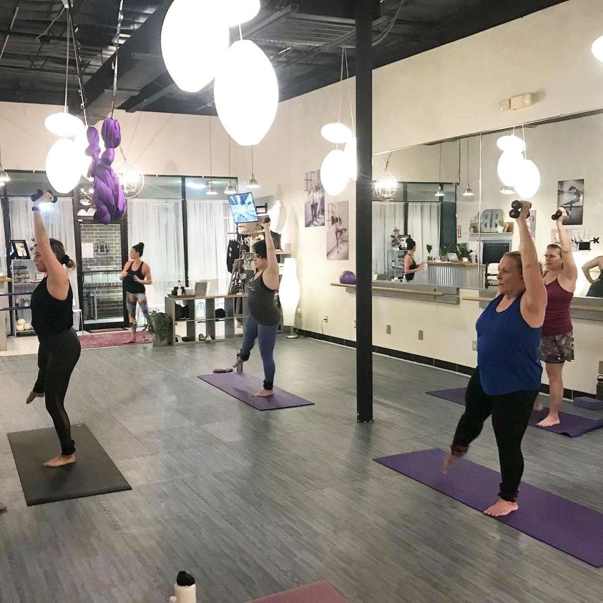 A group of four women is performing a workout routine with weights on yoga mats in a well-lit studio.