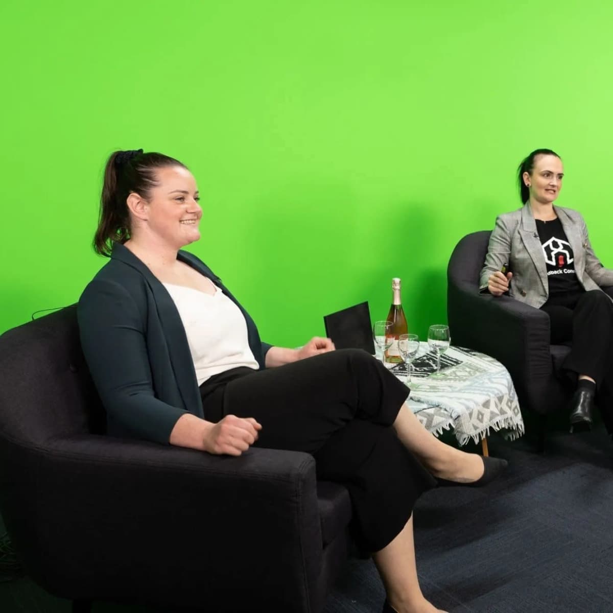 Three individuals are seated on black chairs in front of a bright green screen, with a small table and glasses of champagne beside them.