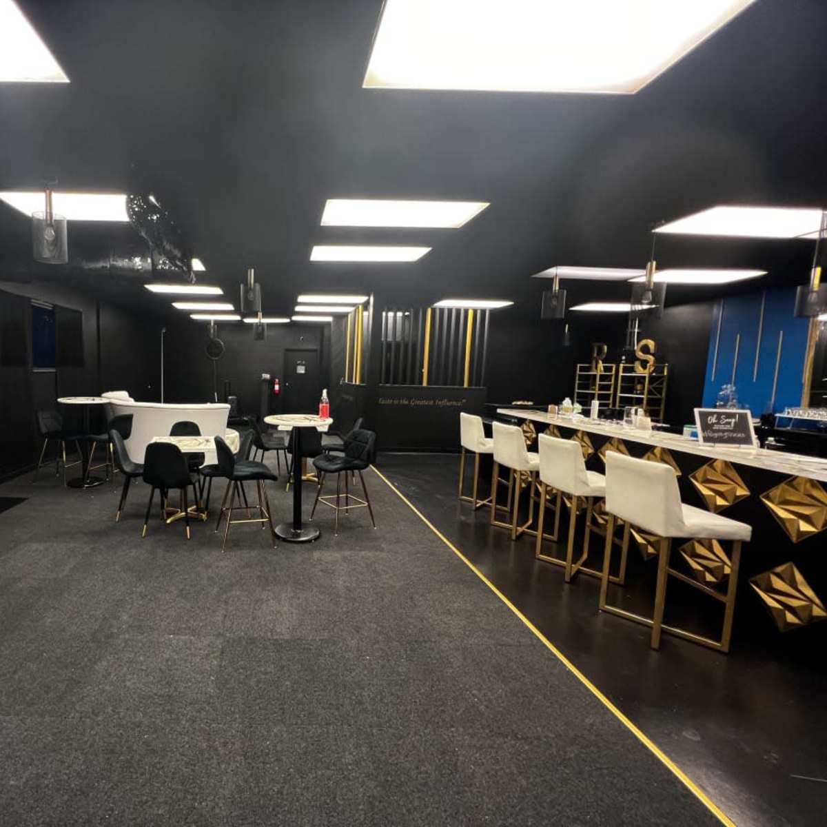 A modern, black-walled interior space featuring a mix of tables and chairs, a bar area, and decorative elements like plants and lighting fixtures.