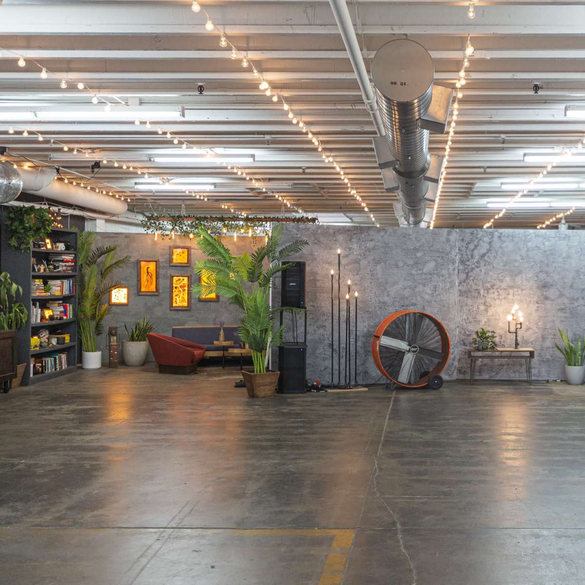 A spacious indoor event area with a gray backdrop, various potted plants, decorative lights, and audio equipment arranged throughout.