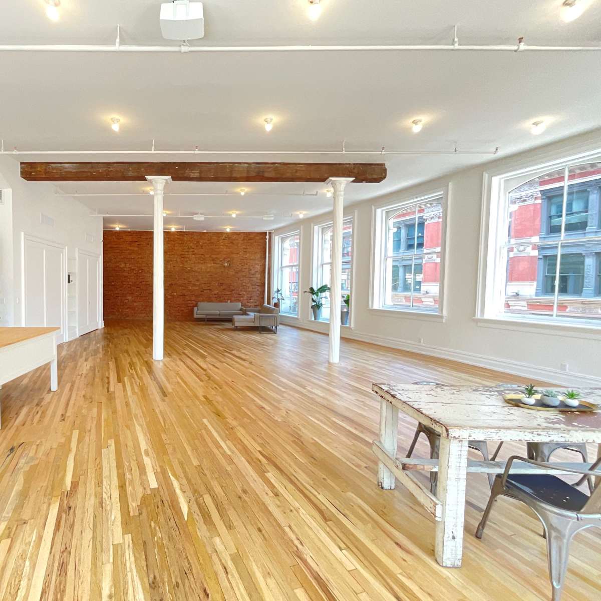 The spacious interior features hardwood floors, exposed brick walls, and large windows, with a kitchen area on one side and a seating area on the other.
