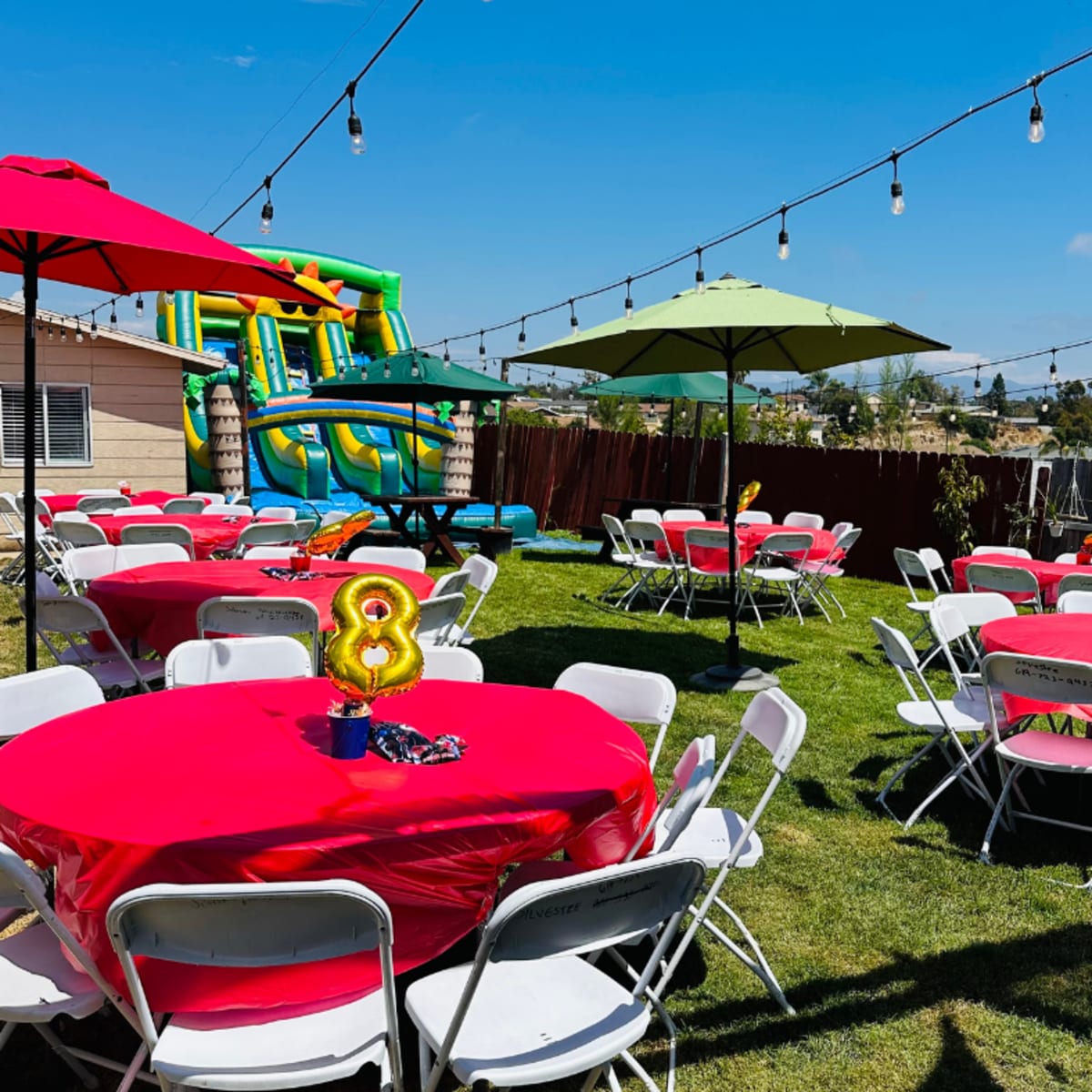 The scene features a backyard party setup with round tables covered in bright pink tablecloths, umbrellas for shade, and an inflatable slide in the background.