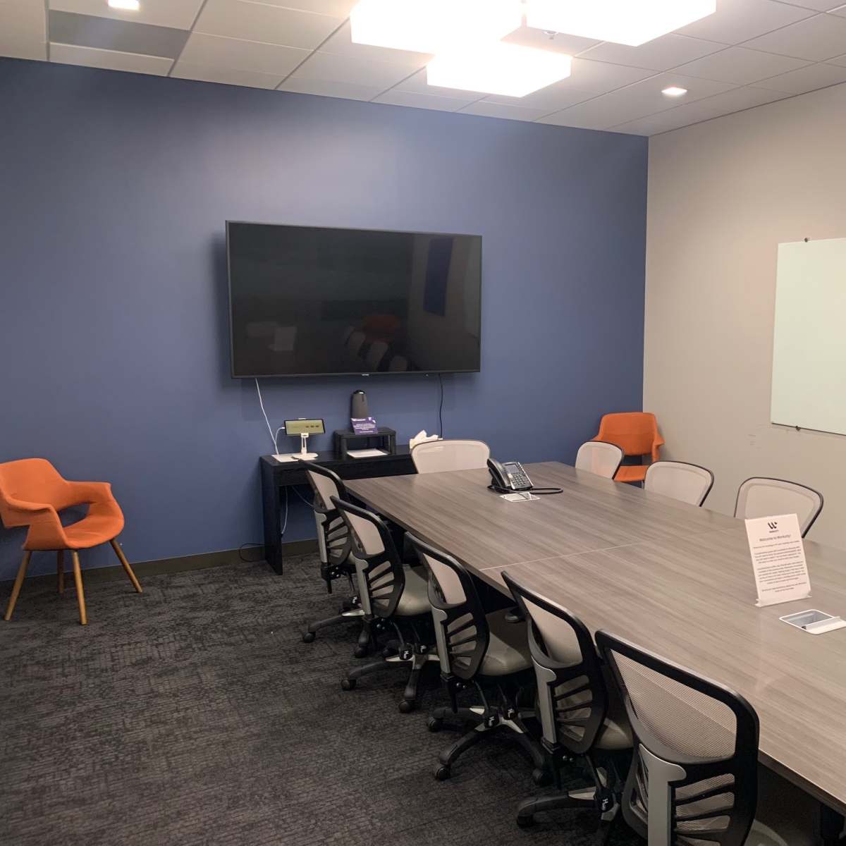 A conference room features a long table surrounded by chairs, a large wall-mounted TV, an orange armchair in the corner, and a whiteboard.