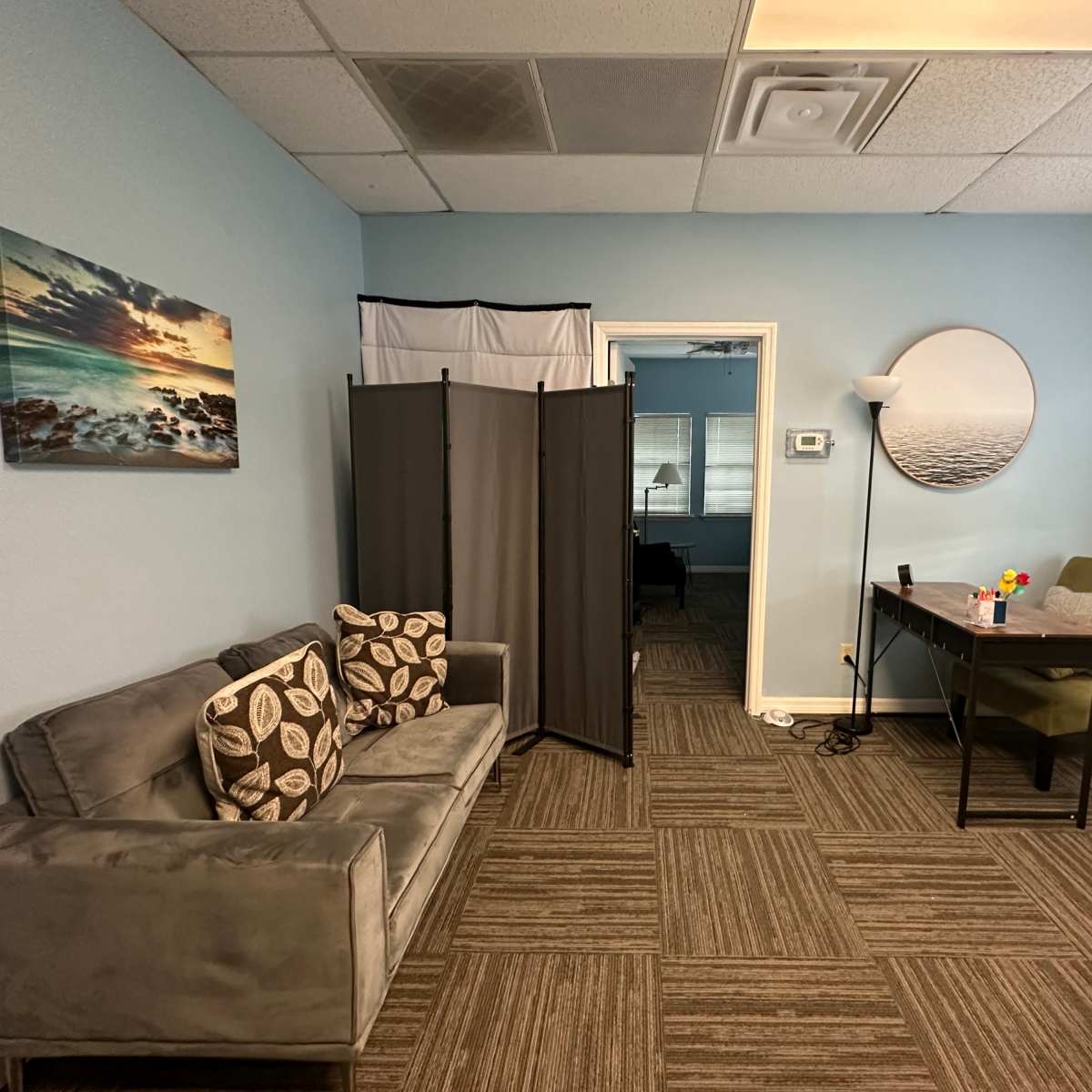 A light blue room featuring a gray couch with patterned pillows, a folding screen, a table with green chairs, and framed artwork on the walls.