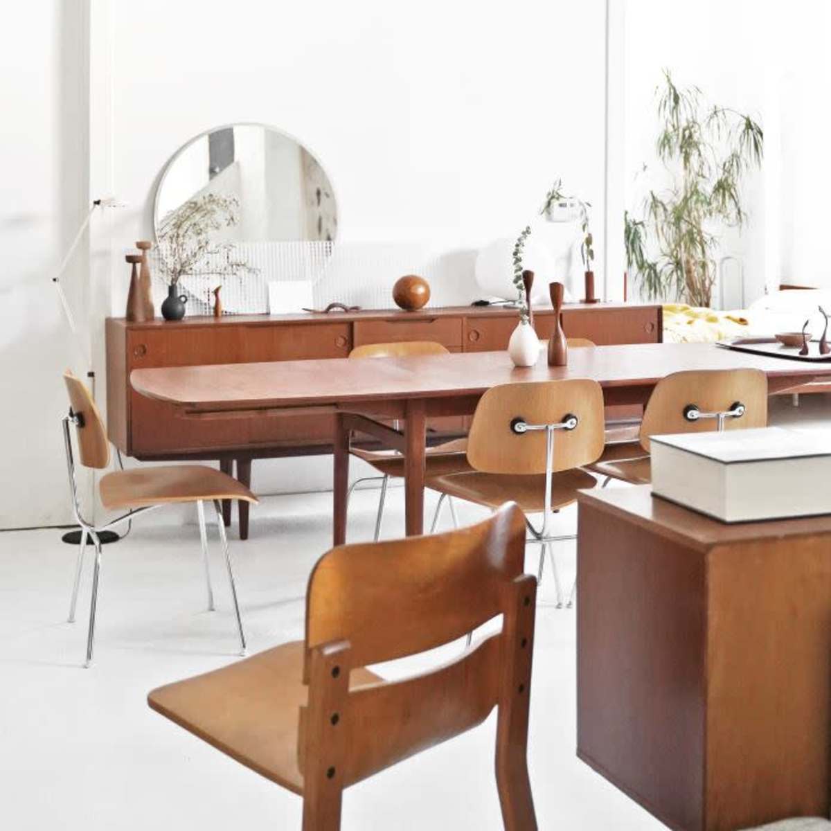 A bright, minimalist interior featuring a wooden dining table surrounded by chairs, with a large mirror and various decorative items on display.