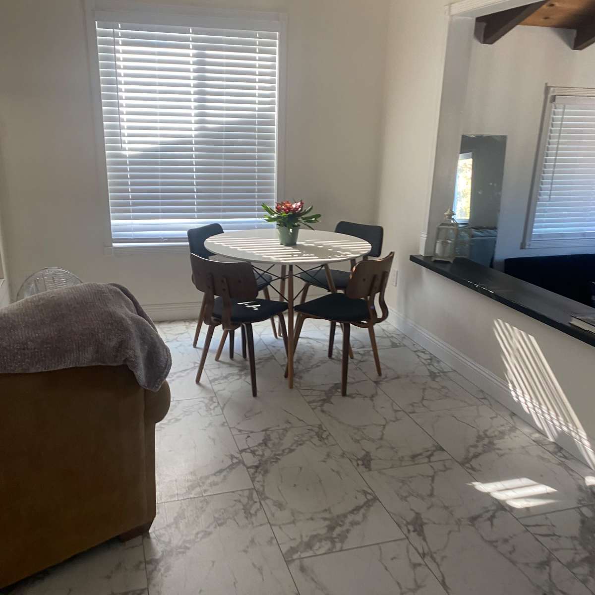 A round dining table with four black chairs is positioned in a bright room featuring tiled flooring and a window with blinds.