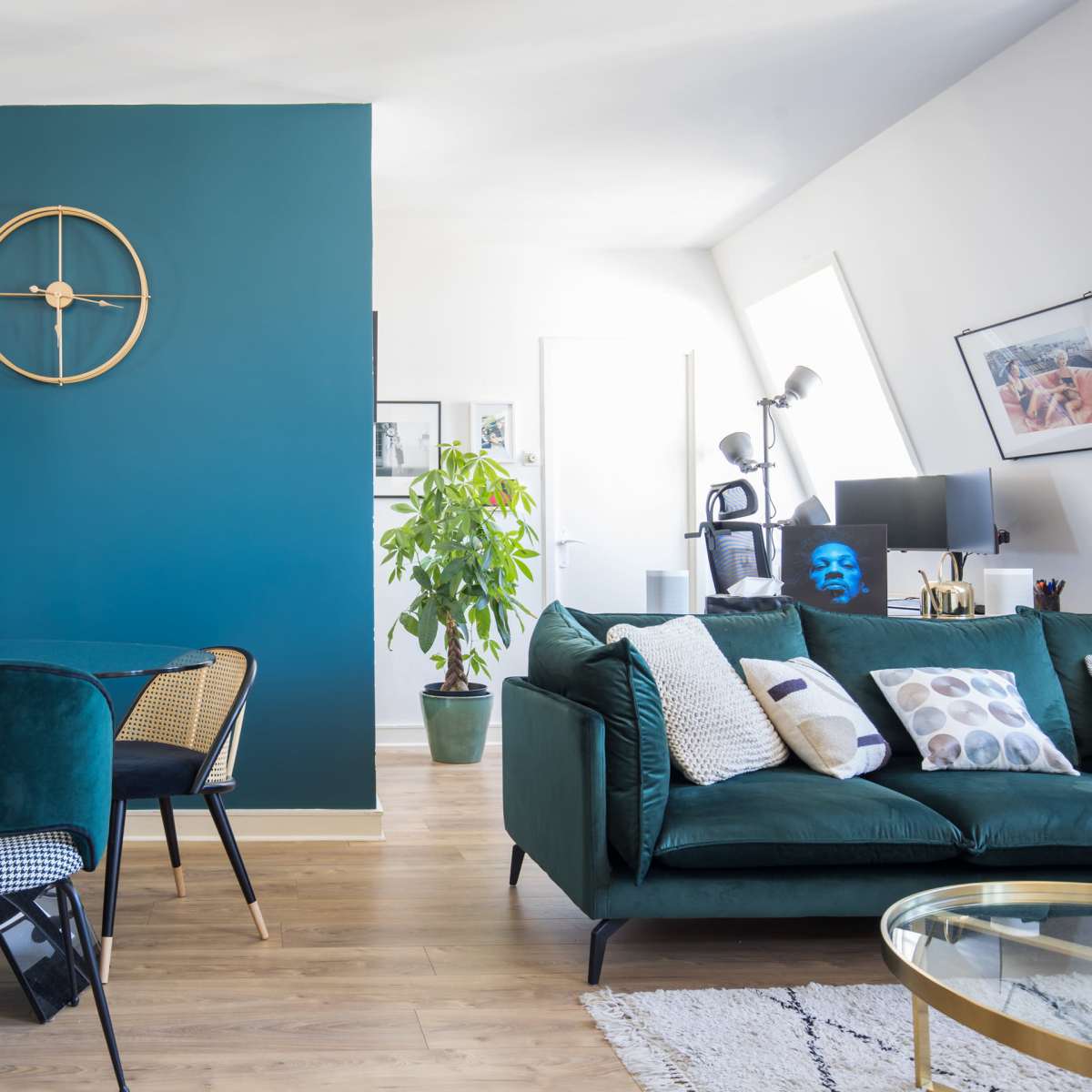 A modern living space features a green accent wall, a circular clock, a dining table with mixed seating, and a potted plant, alongside a cozy sofa adorned with decorative pillows.