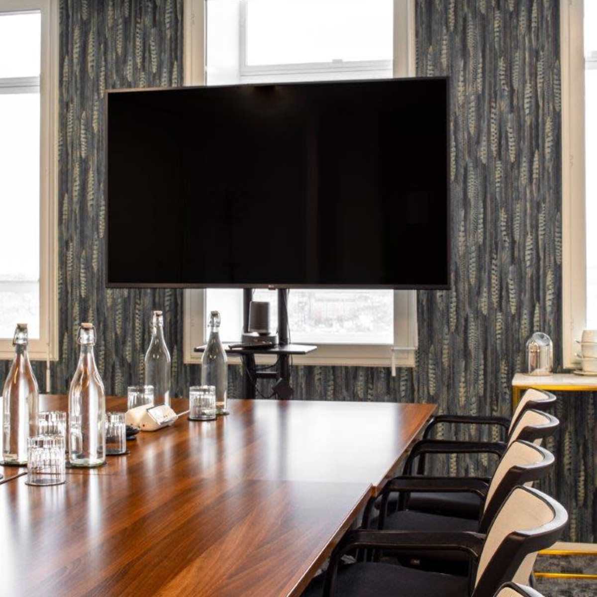 A modern conference room features a long wooden table, black chairs, a large mounted television, and windows with patterned wallpaper.