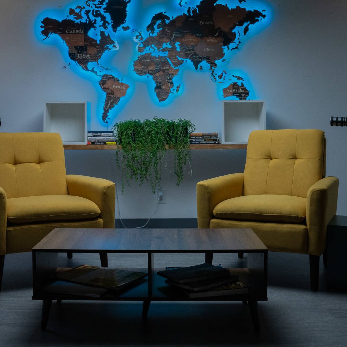 Two yellow chairs facing each other in a room with a world map illuminated in blue on the wall behind them, accompanied by a coffee table and a hanging plant.