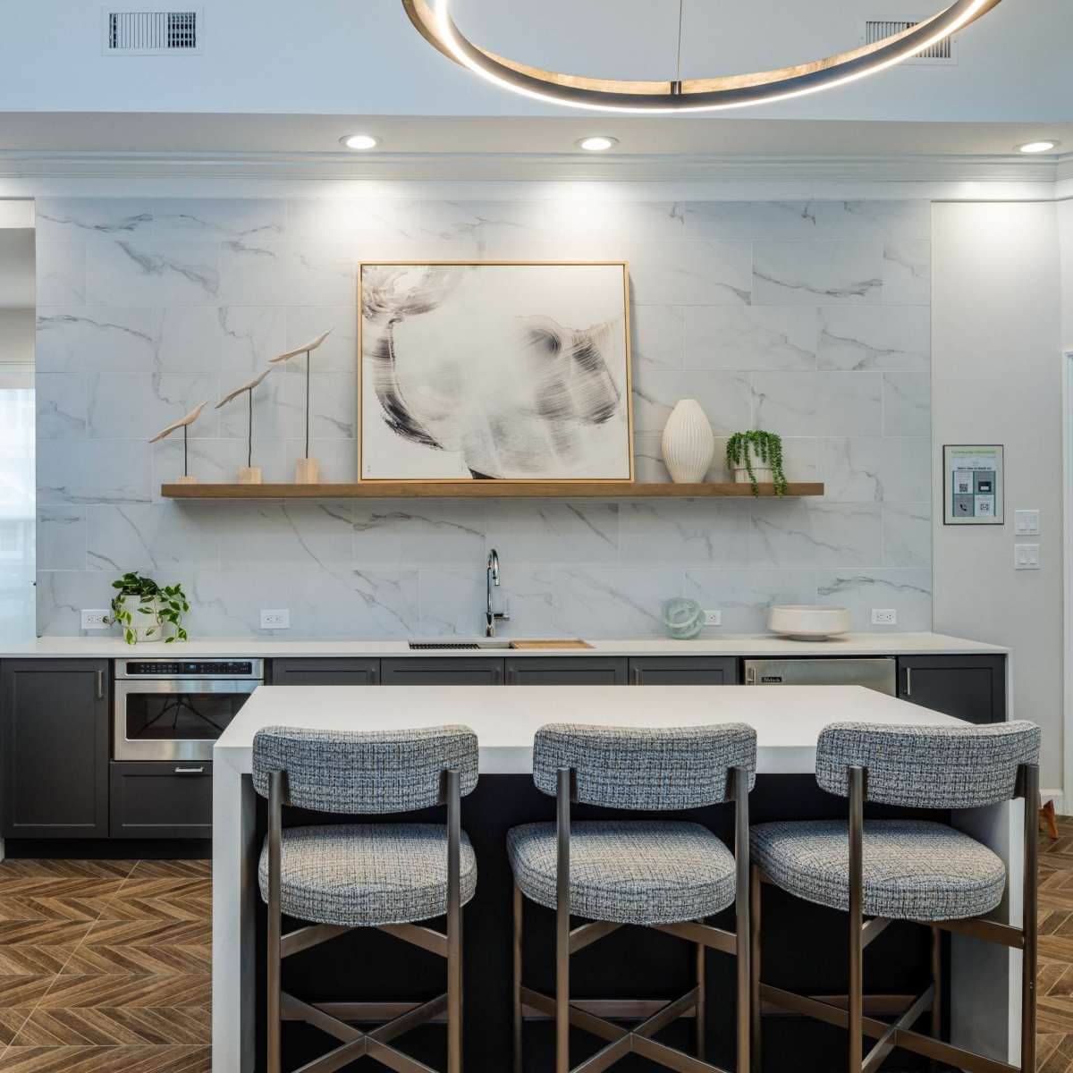 A modern kitchen with gray cabinetry, a white island bar with three upholstered stools, and a marble accent wall featuring a piece of artwork and a small shelf with a plant.