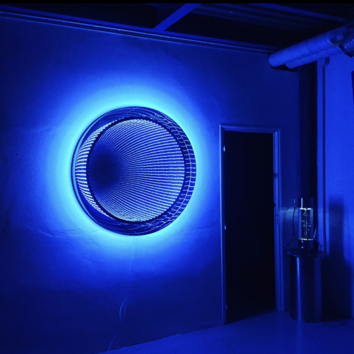 A circular, wire mesh art piece is illuminated with blue light, mounted on a wall beside an open door in a dimly lit room.
