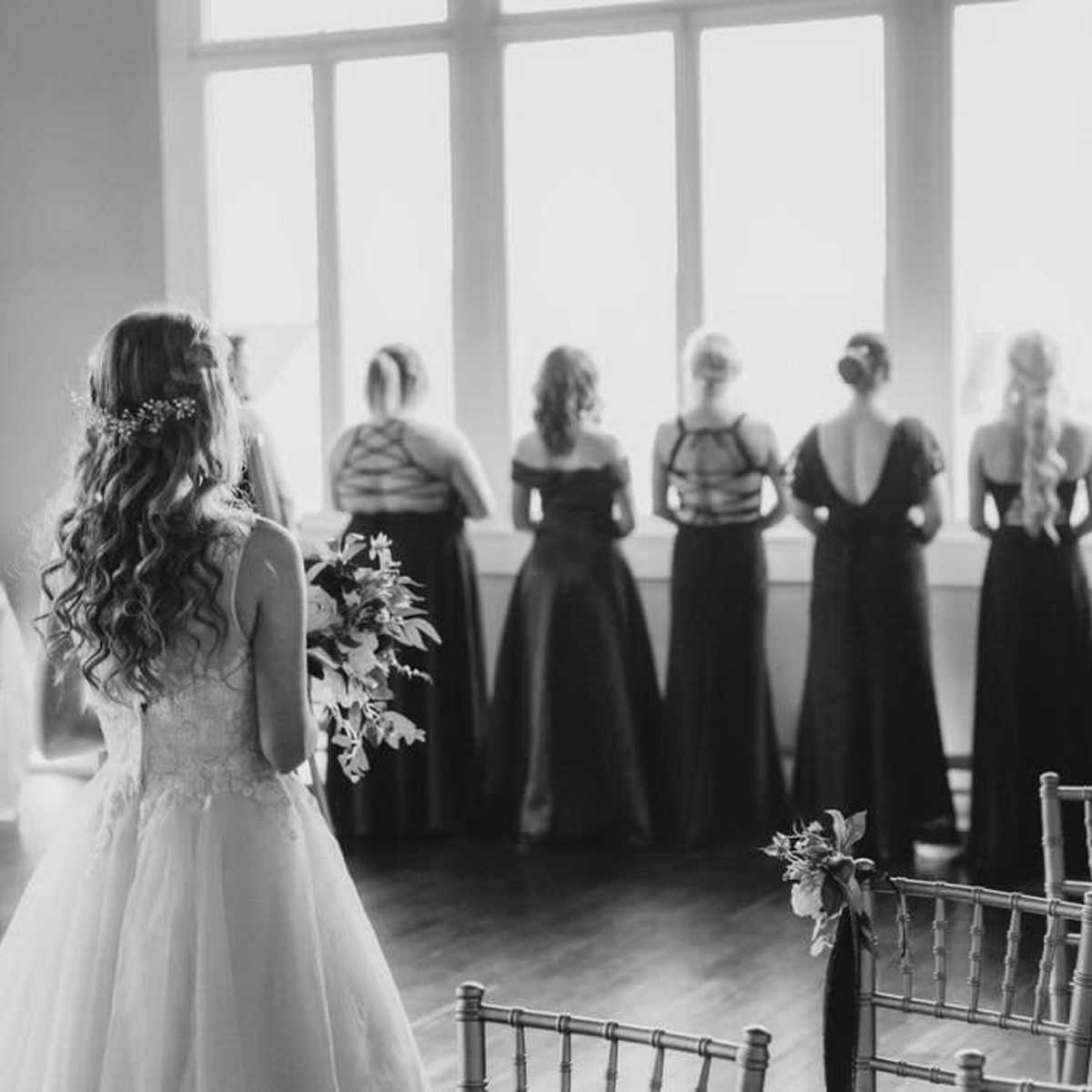 A bride in a white dress stands with her back to the camera, while her bridesmaids in black dresses gaze out the window.