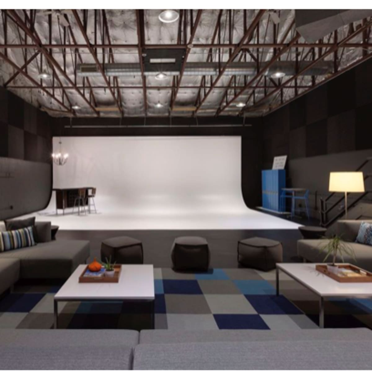 A spacious studio with a white backdrop, surrounded by modern furniture including a sectional sofa, coffee tables, and individual seating, under a structured ceiling.