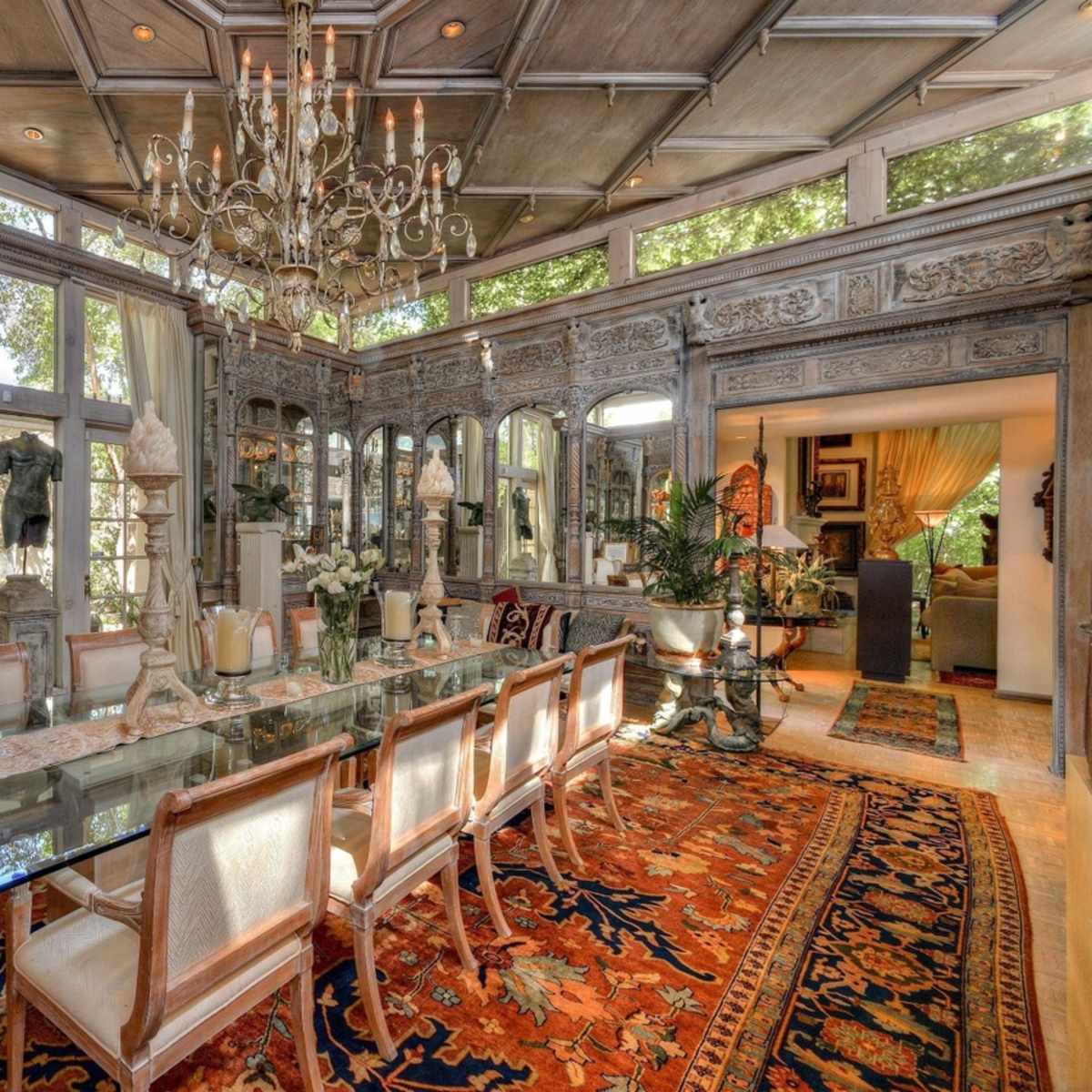 An elegant dining room featuring a large glass table surrounded by upholstered chairs, ornate wooden detailing on the walls, a chandelier, and large windows providing natural light.