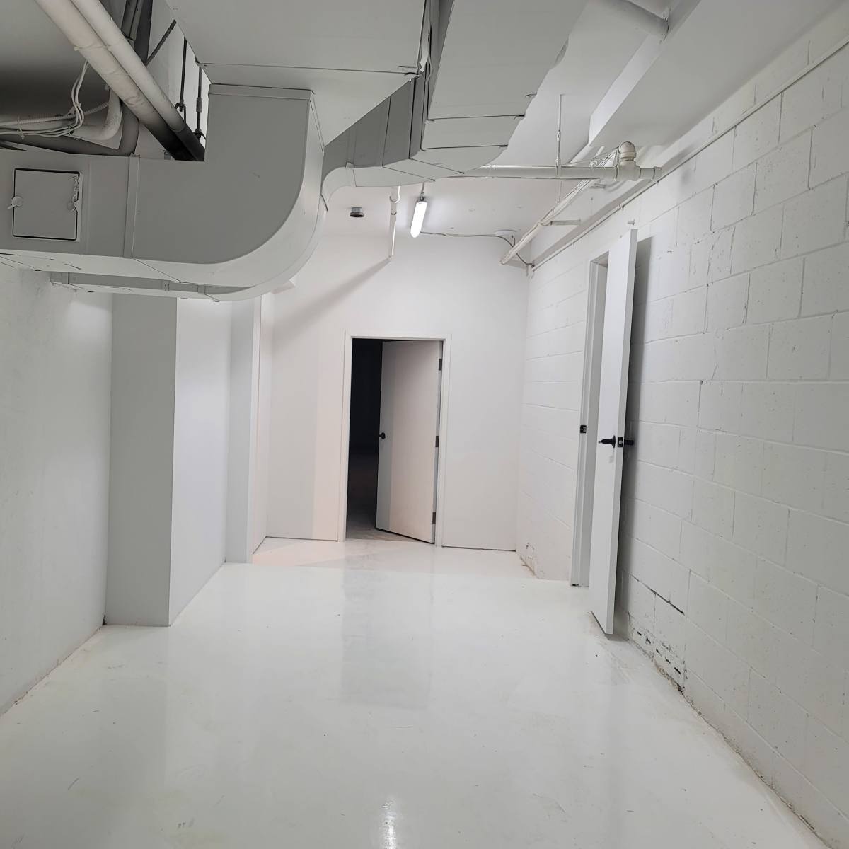 A light-colored corridor in a basement with several doors and exposed ductwork on the ceiling.