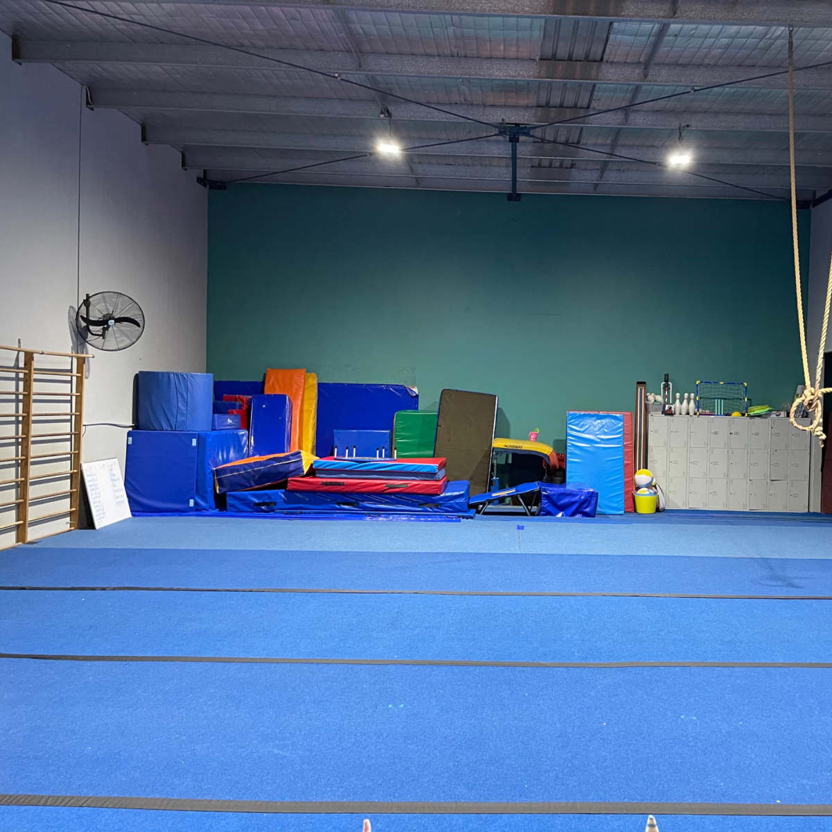 A gymnasium floor covered in blue mats, surrounded by various gym equipment, including colorful mats and a climbing rope.
